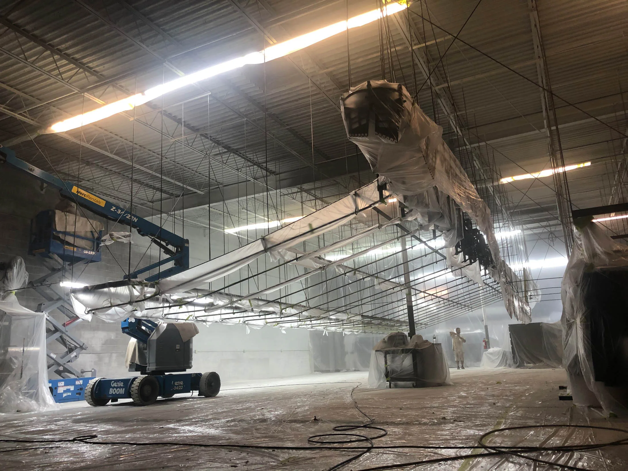 Commercial painting site with boom lift, workers in protective gear, and covered surfaces in preparation for professional painting services in a spacious interior.