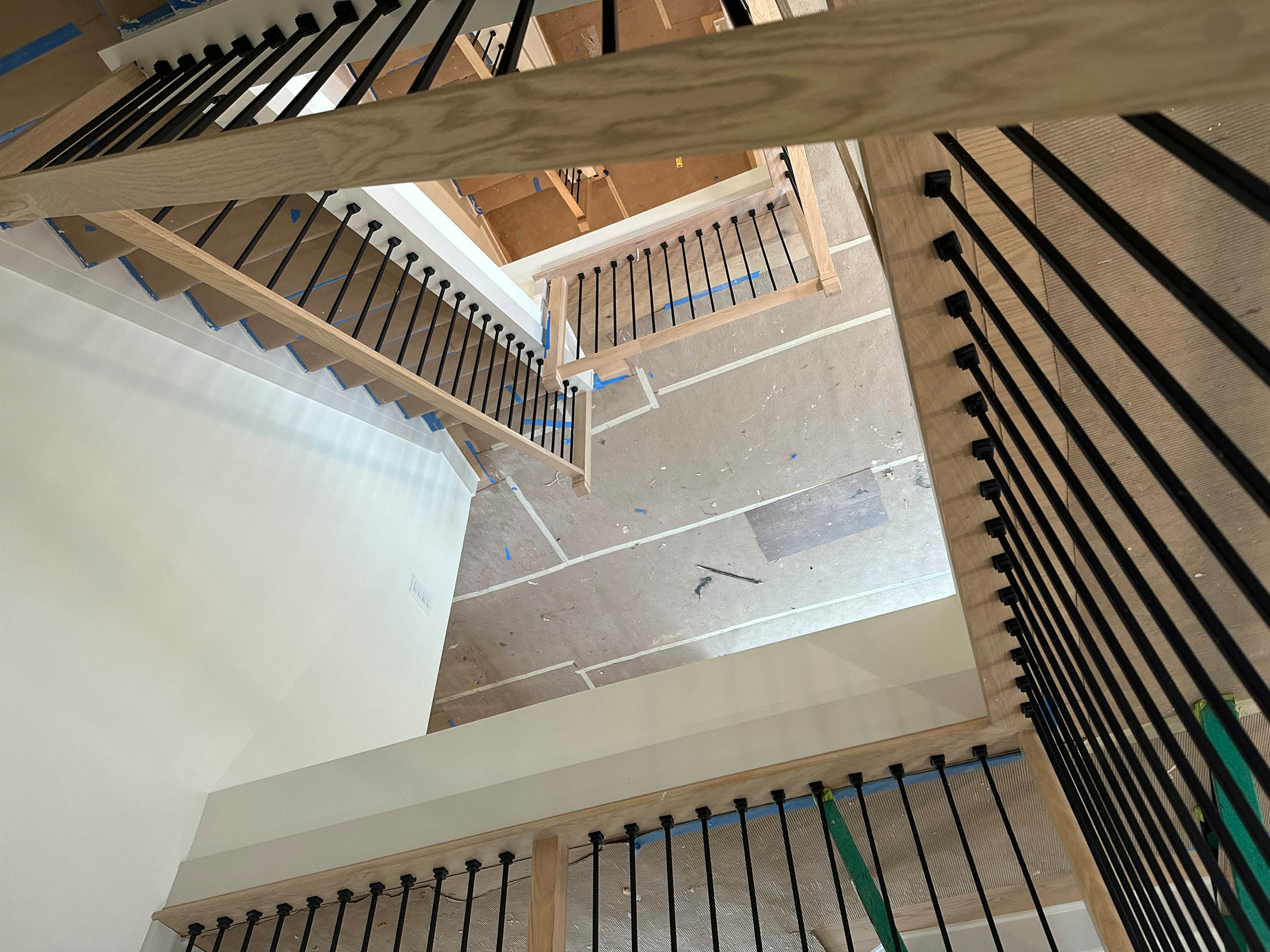 View looking down a multi-story stairwell with black metal railings and wood handrails; floors below are covered with protective paper during construction.