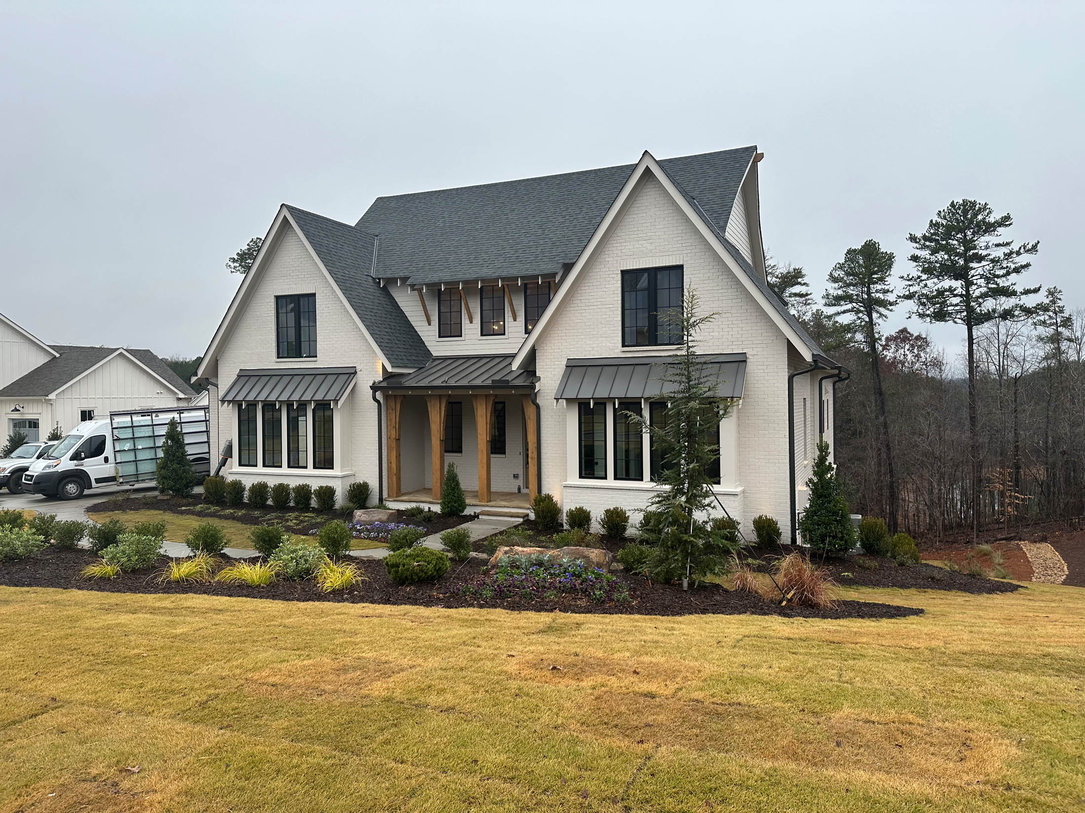 A modern two-story house with light-colored brick, dark roof, and multiple windows, surrounded by landscaping and a neatly maintained lawn on a cloudy day.