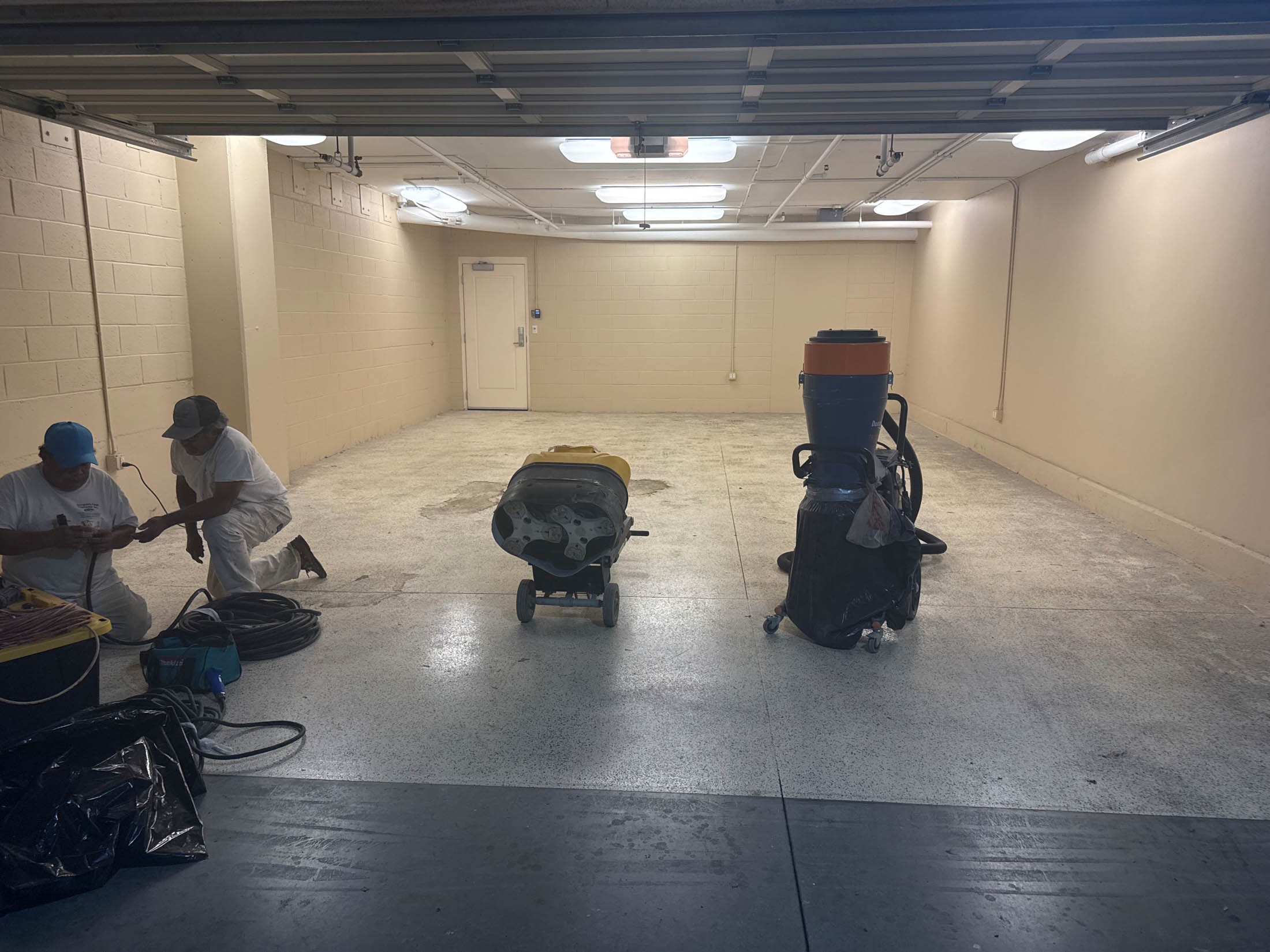 Two workers clean and prepare an empty garage with cleaning equipment and supplies placed on the floor under fluorescent lighting.