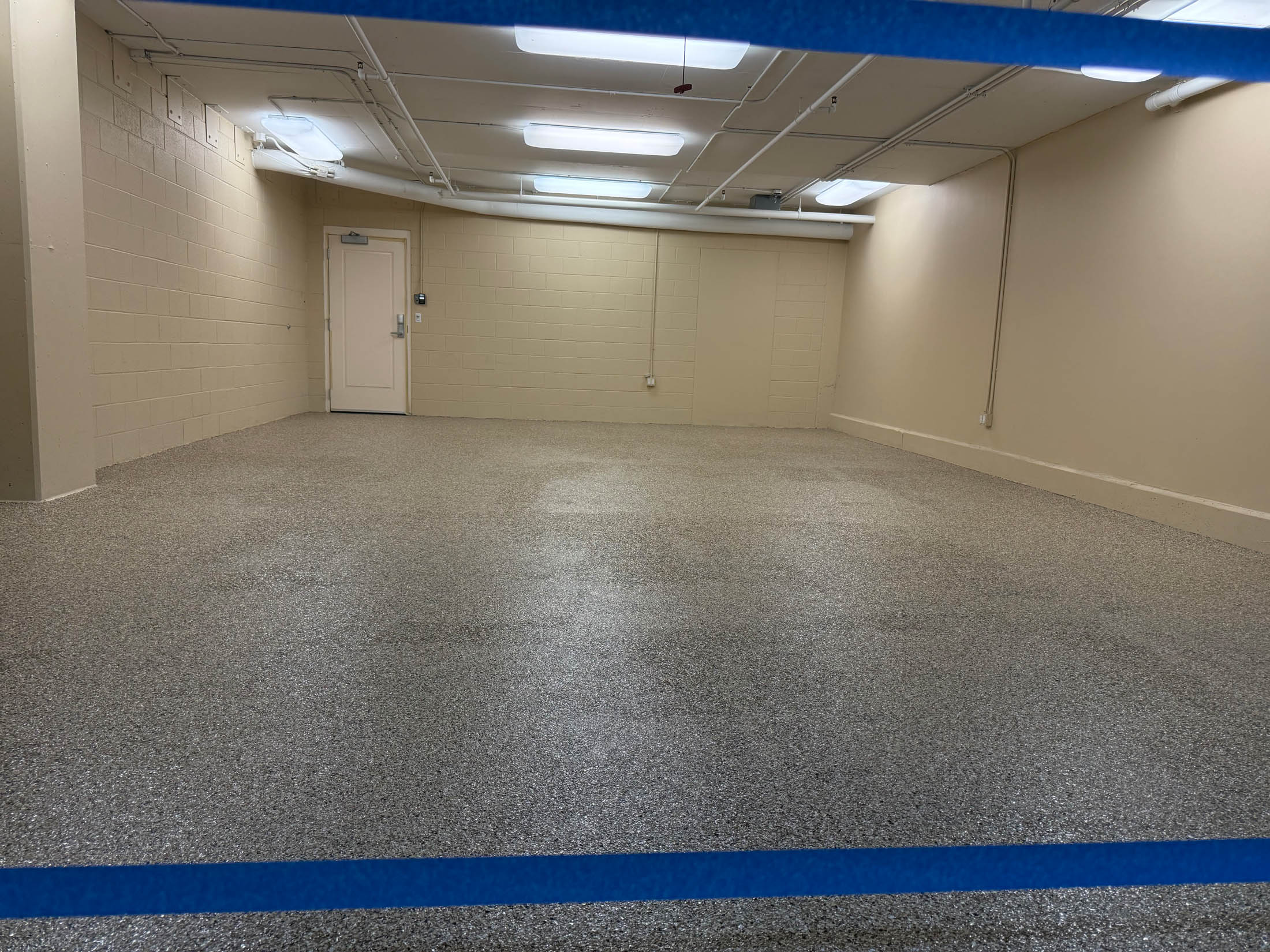 Empty, well-lit room with beige walls, a speckled gray floor, fluorescent ceiling lights, and a closed white door in the back wall. Blue tape runs horizontally in the foreground.