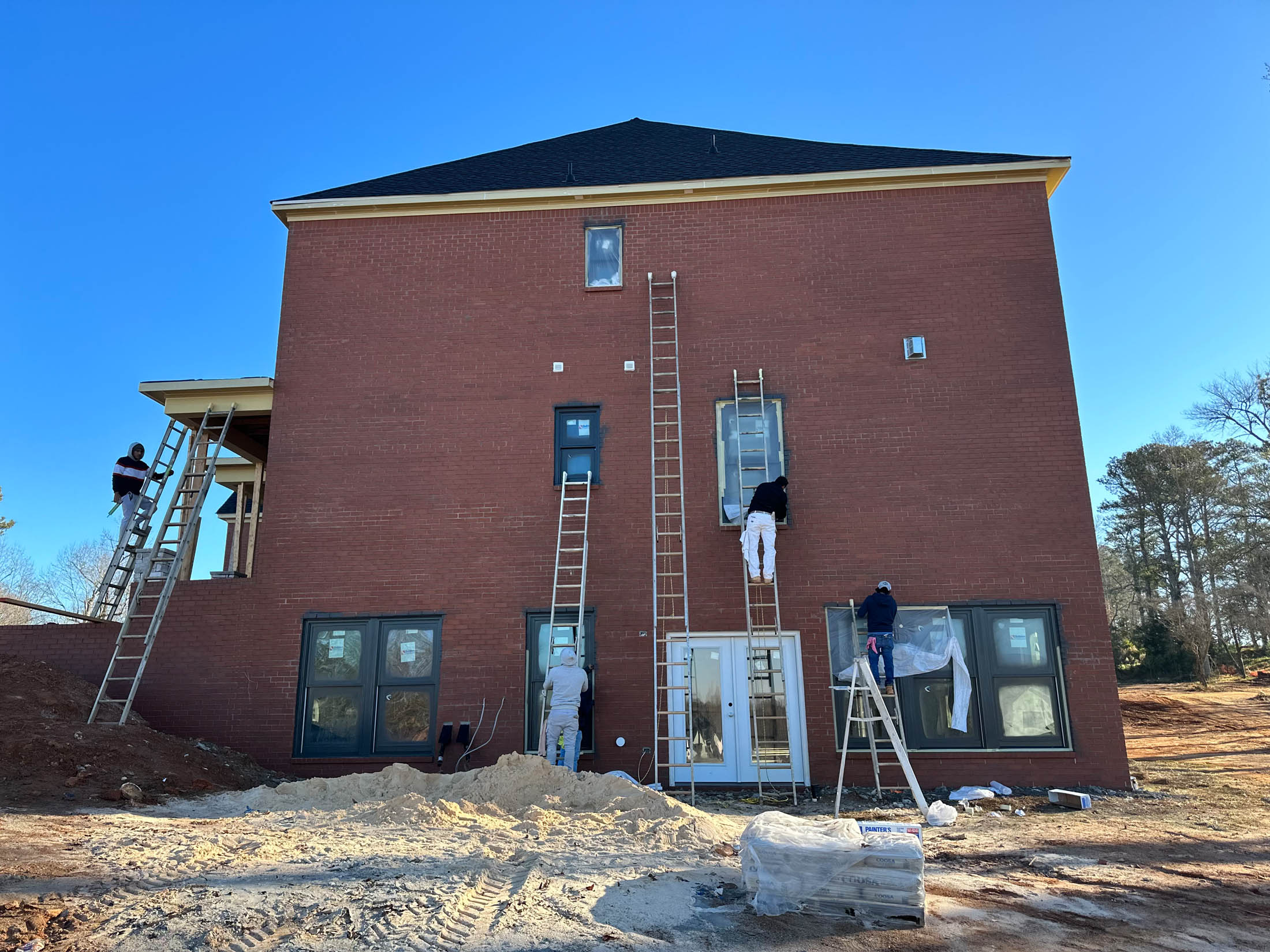 Four workers on ladders are painting or working on the exterior brick wall of a two-story house on a clear day. Construction materials are on the ground.