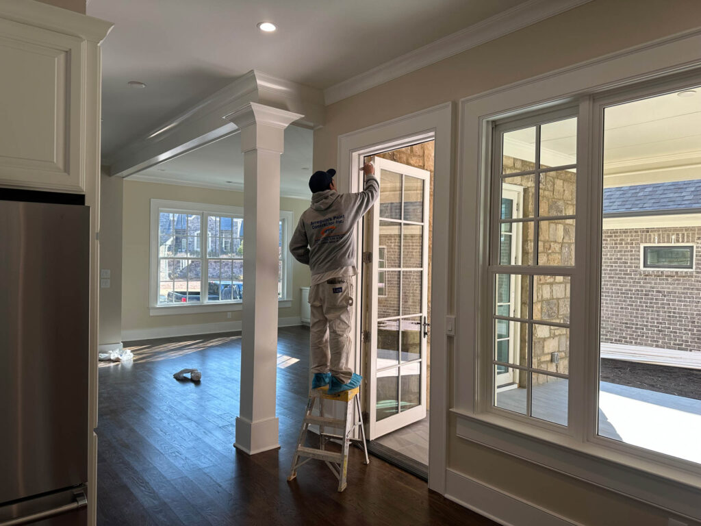 A painter standing on a small ladder applies paint to a glass patio door frame in a bright, empty room. Contact Arreguin’s Painting for expert service.