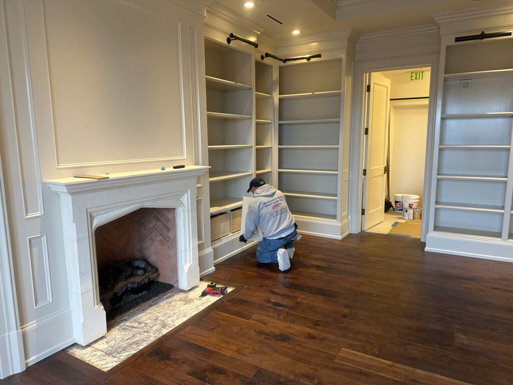 A person kneels on a hardwood floor installing shelves in a built-in bookcase next to a fireplace in a room under renovation.