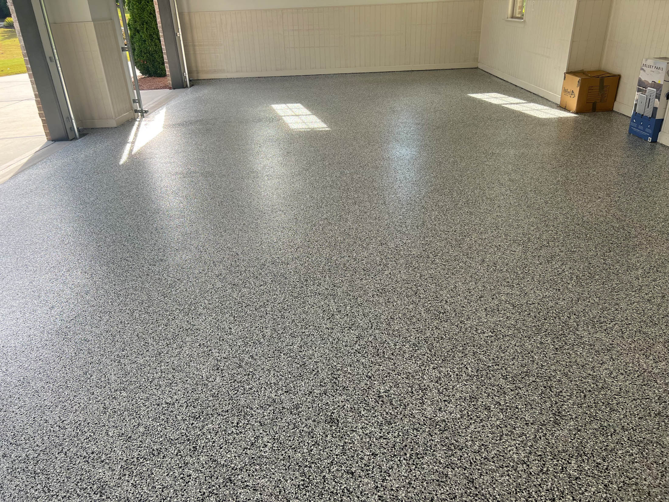 A clean, empty garage with a gray speckled epoxy floor, white walls, open doors, and cardboard boxes in the corner.