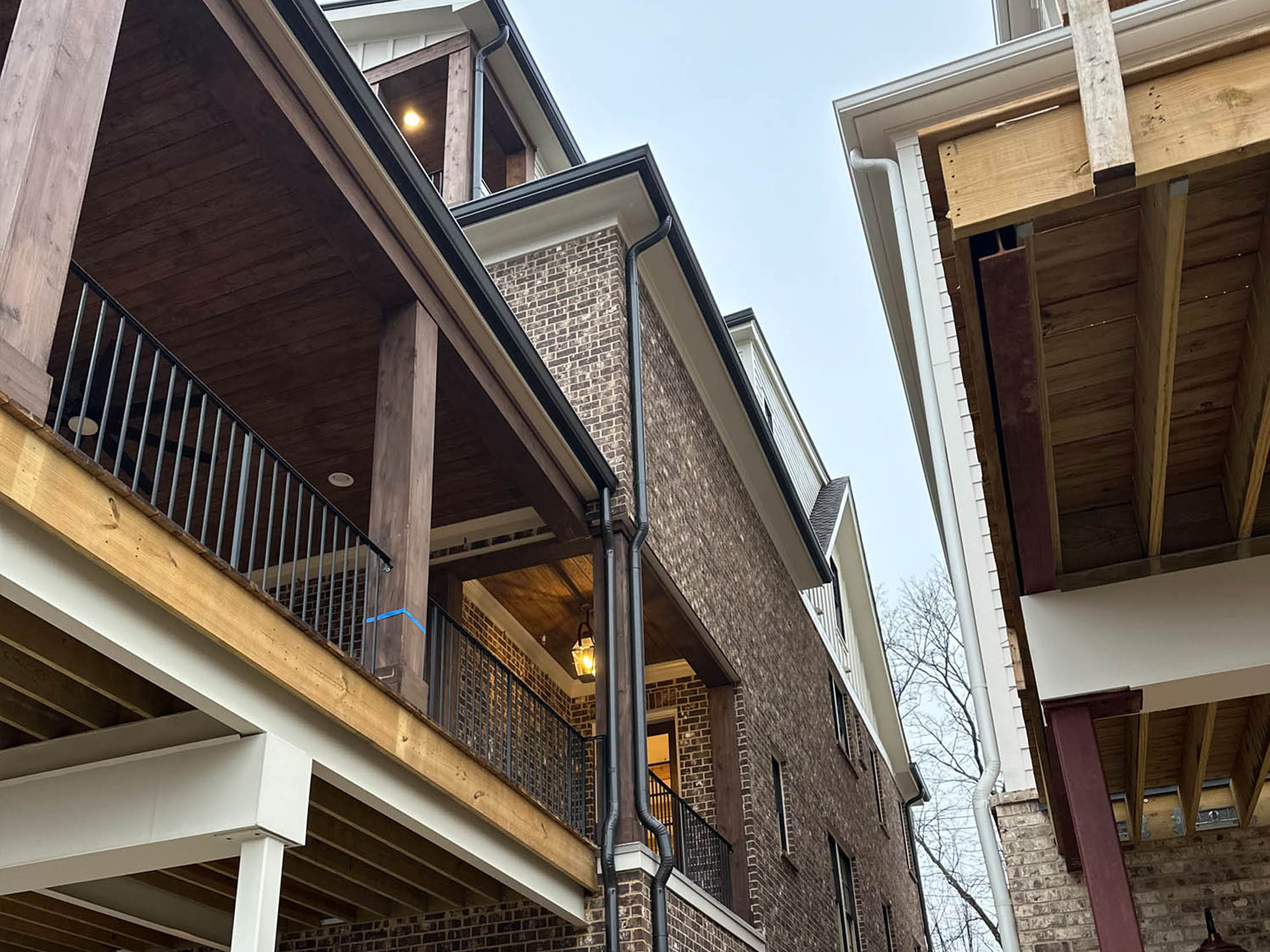 View looking up at two multi-story brick houses with covered balconies, wooden beams, and exposed rafters under an overcast sky.