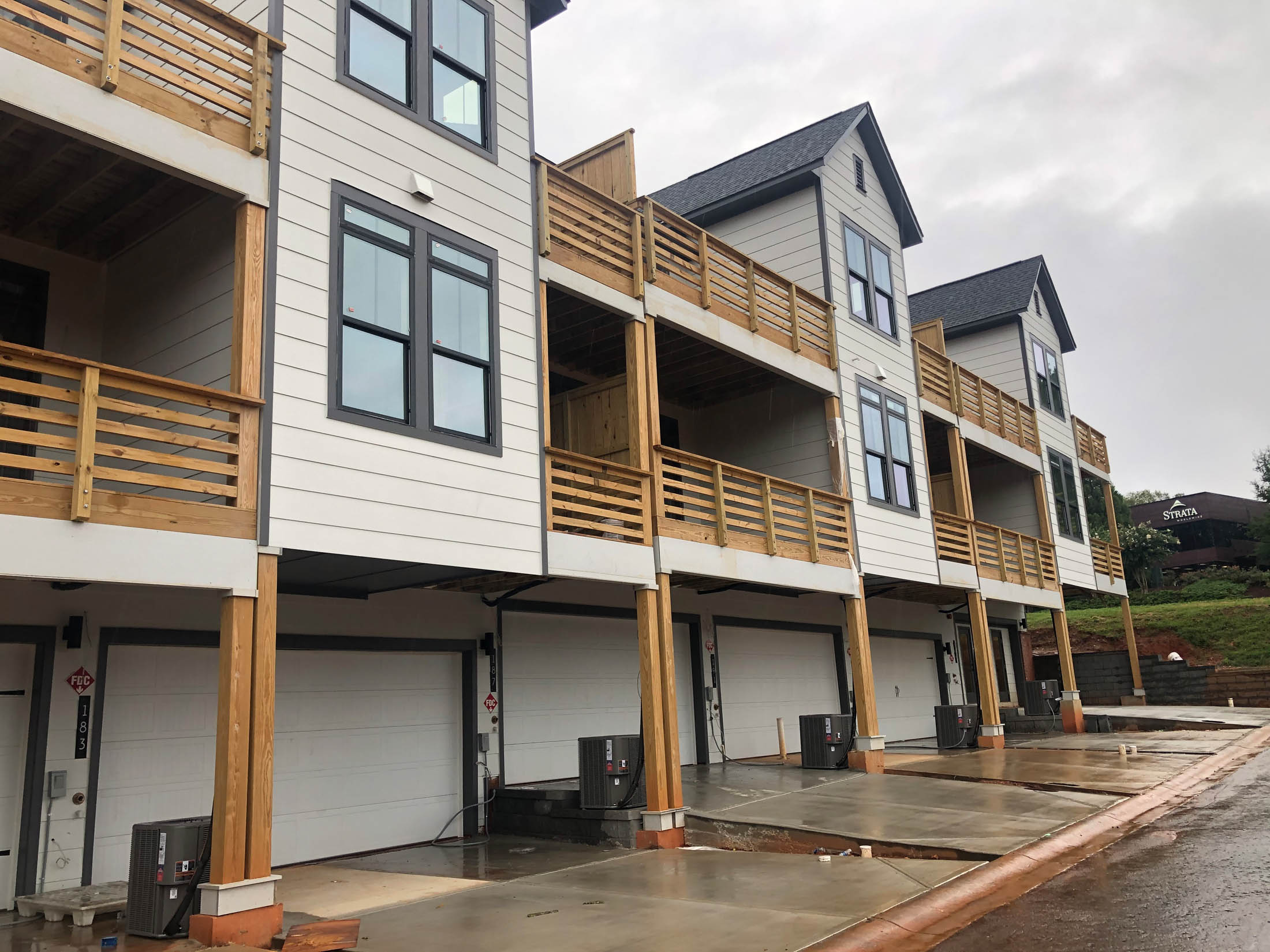 Row of modern three-story townhouses with wooden balconies, double garage doors, and concrete driveways on a cloudy day.