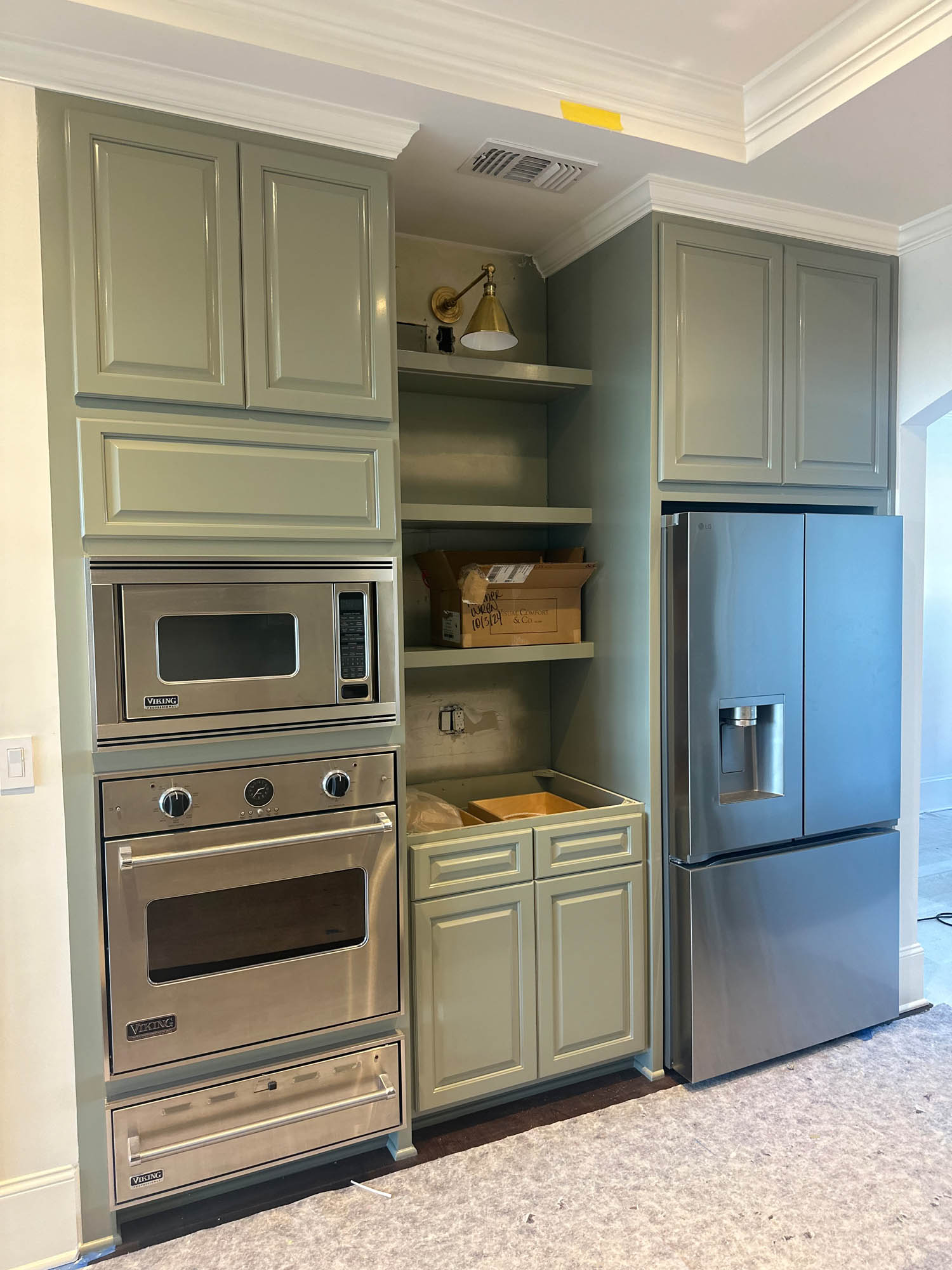 Built-in kitchen area with sage green cabinets, stainless steel appliances including a microwave, oven, and refrigerator, and an unfinished open shelving section with exposed wiring.