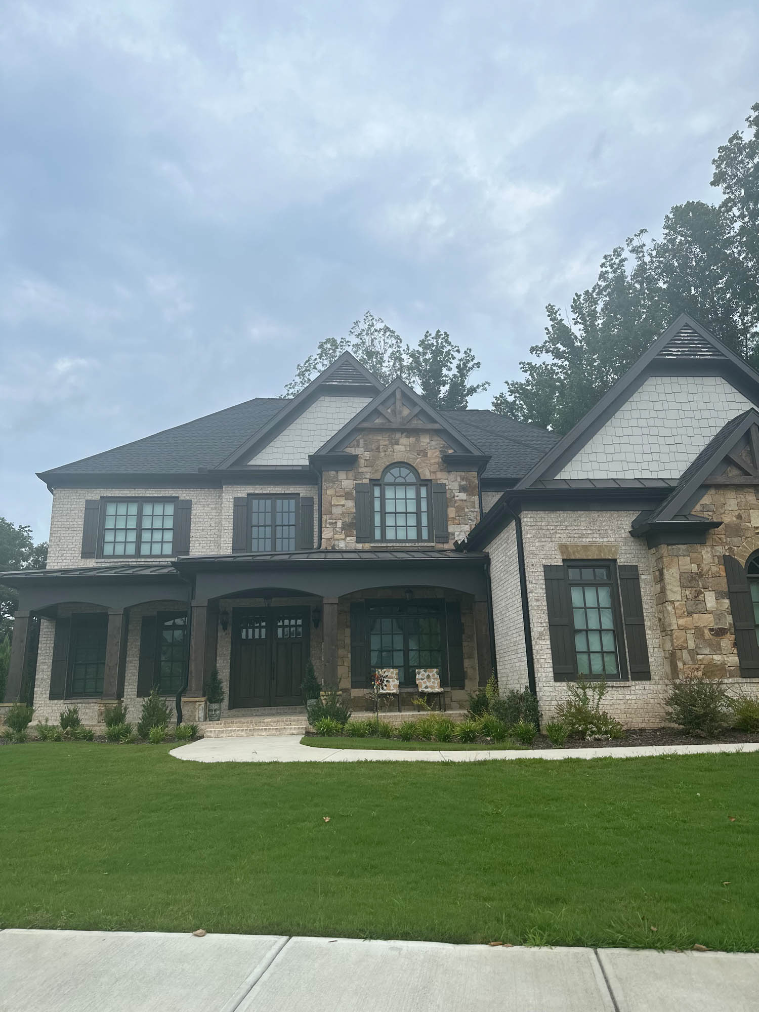 Two-story house with brick and stone exterior, black shutters, and a covered front porch, set on a well-maintained lawn with a curved walkway.