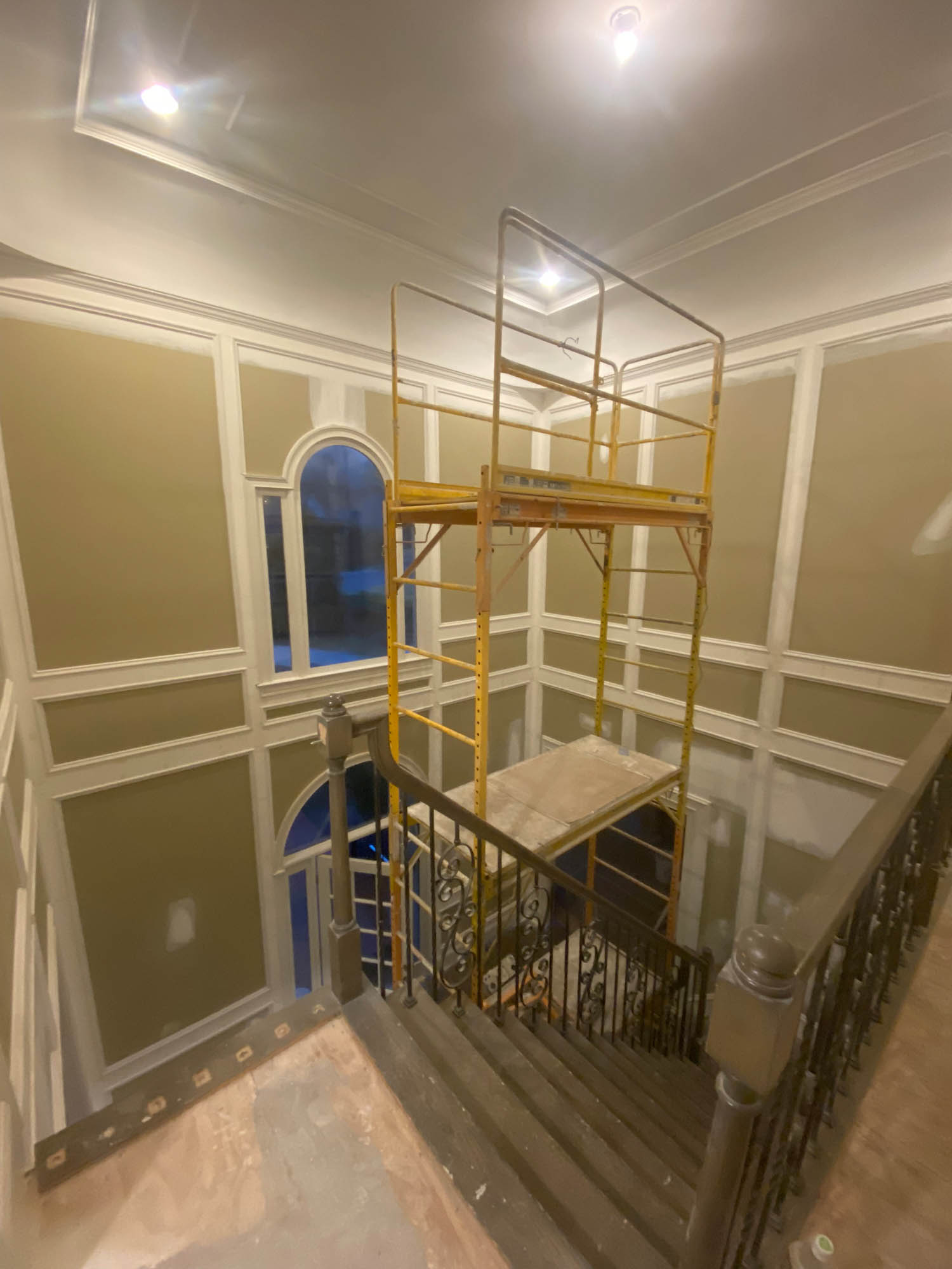 Scaffolding is set up above a staircase landing inside a building with high ceilings, recessed lighting, and paneled walls.