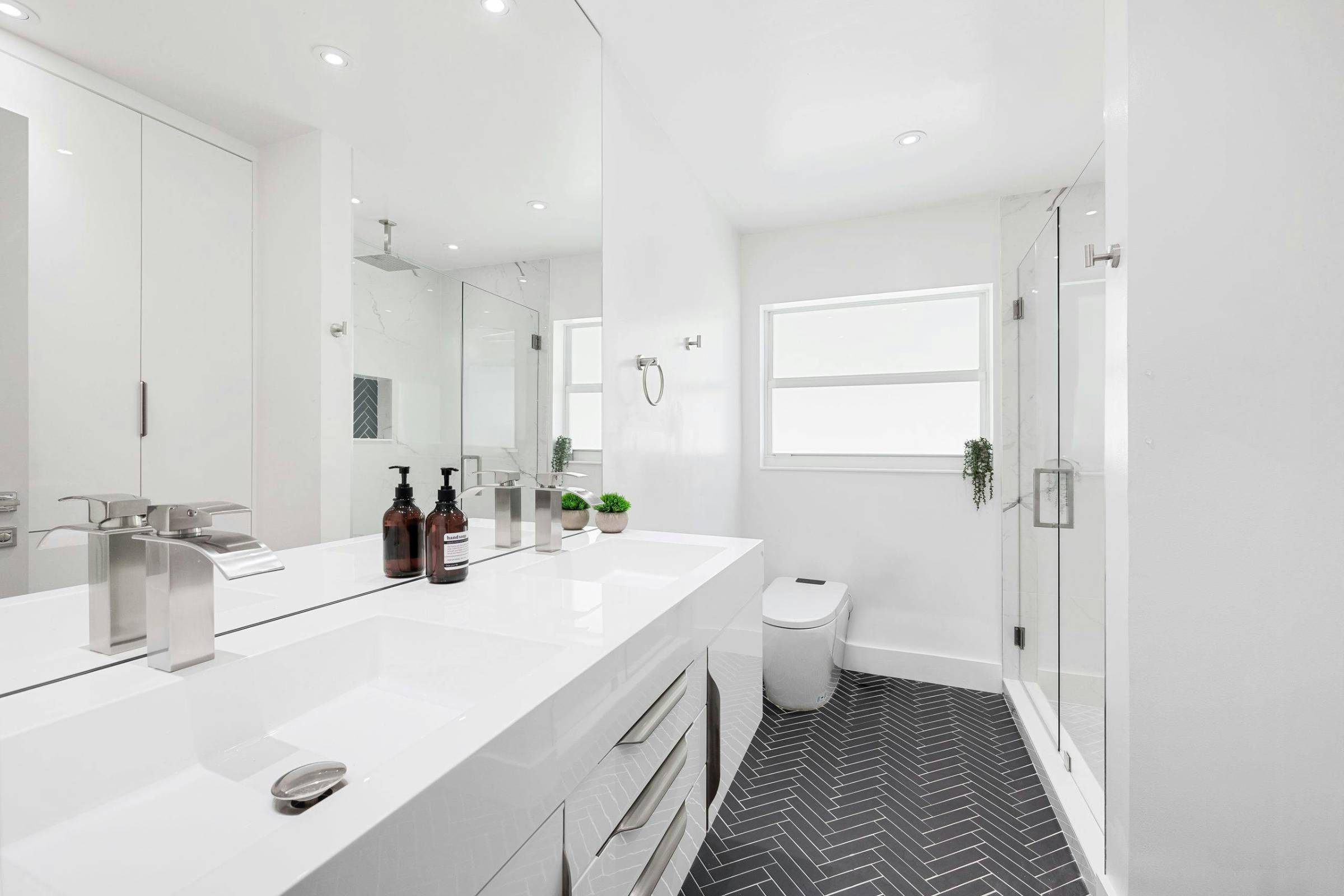 Modern bathroom with a double sink vanity, black herringbone tile floor, glass shower enclosure, and a window letting in natural light—perfectly complemented by fresh New Construction Painting.