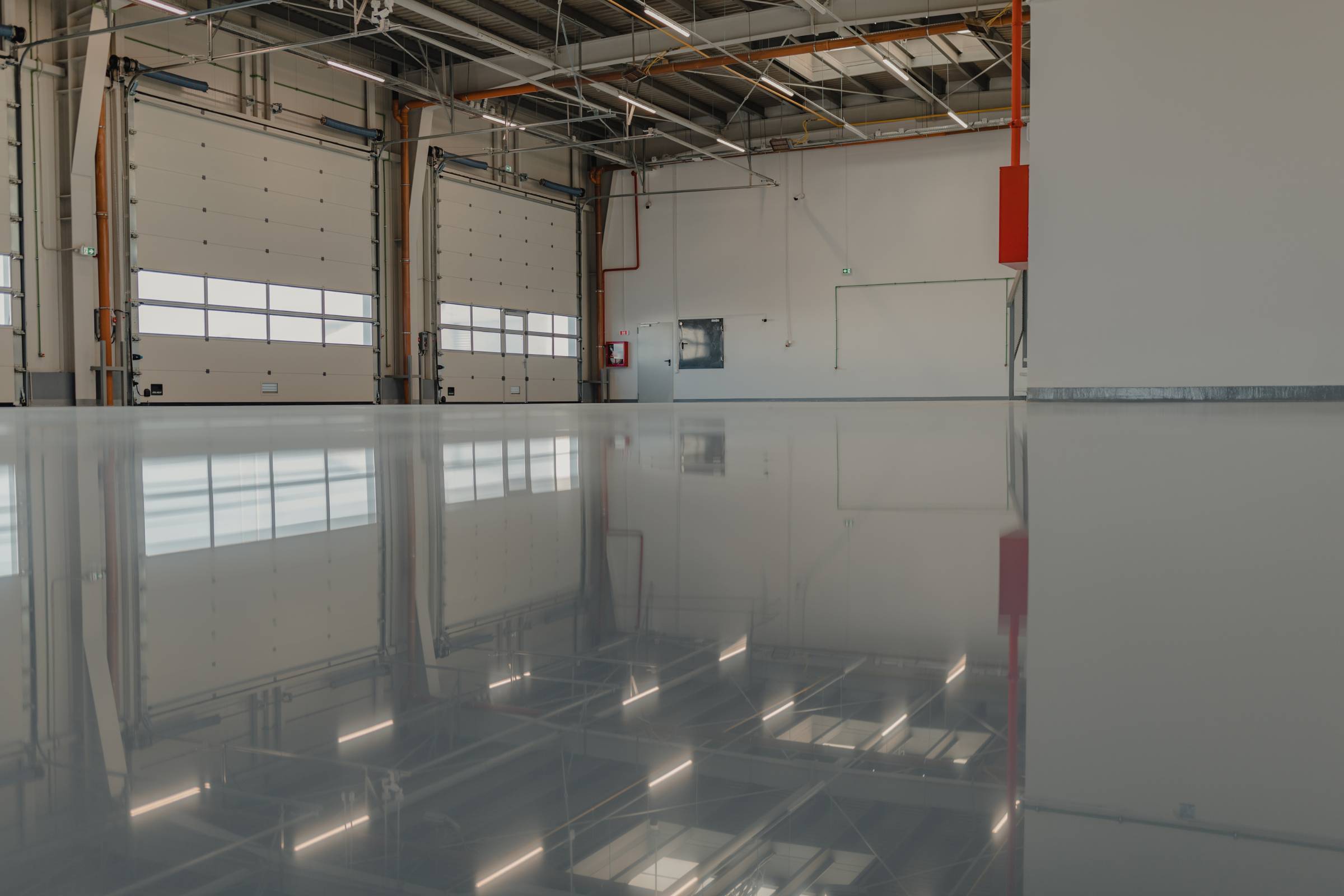 Large empty industrial warehouse with high ceilings, closed overhead doors, exposed pipes, and a polished, reflective concrete floor—ideal for new construction painting projects.