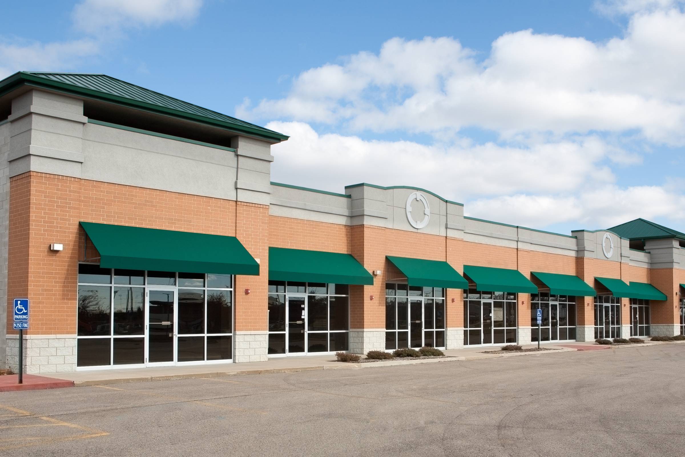 Row of empty storefronts with green awnings in a modern shopping plaza, featuring large glass windows and a mostly empty parking lot—an ideal spot for New Construction Painting or other professional Painting Services under a partly cloudy sky.