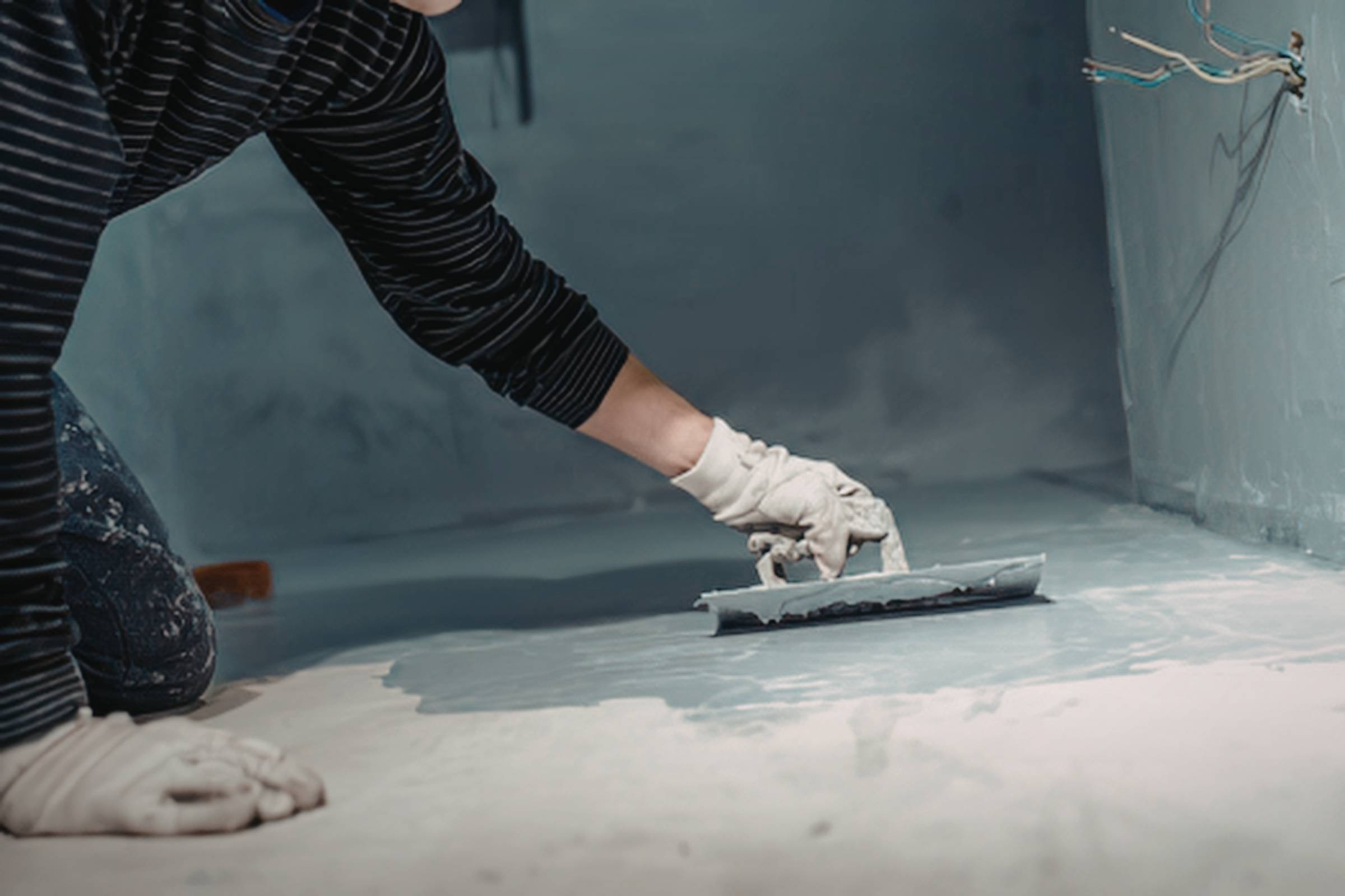 Person wearing gloves applies a smooth layer of material to a floor with a trowel in a room undergoing renovation or New Construction Painting.