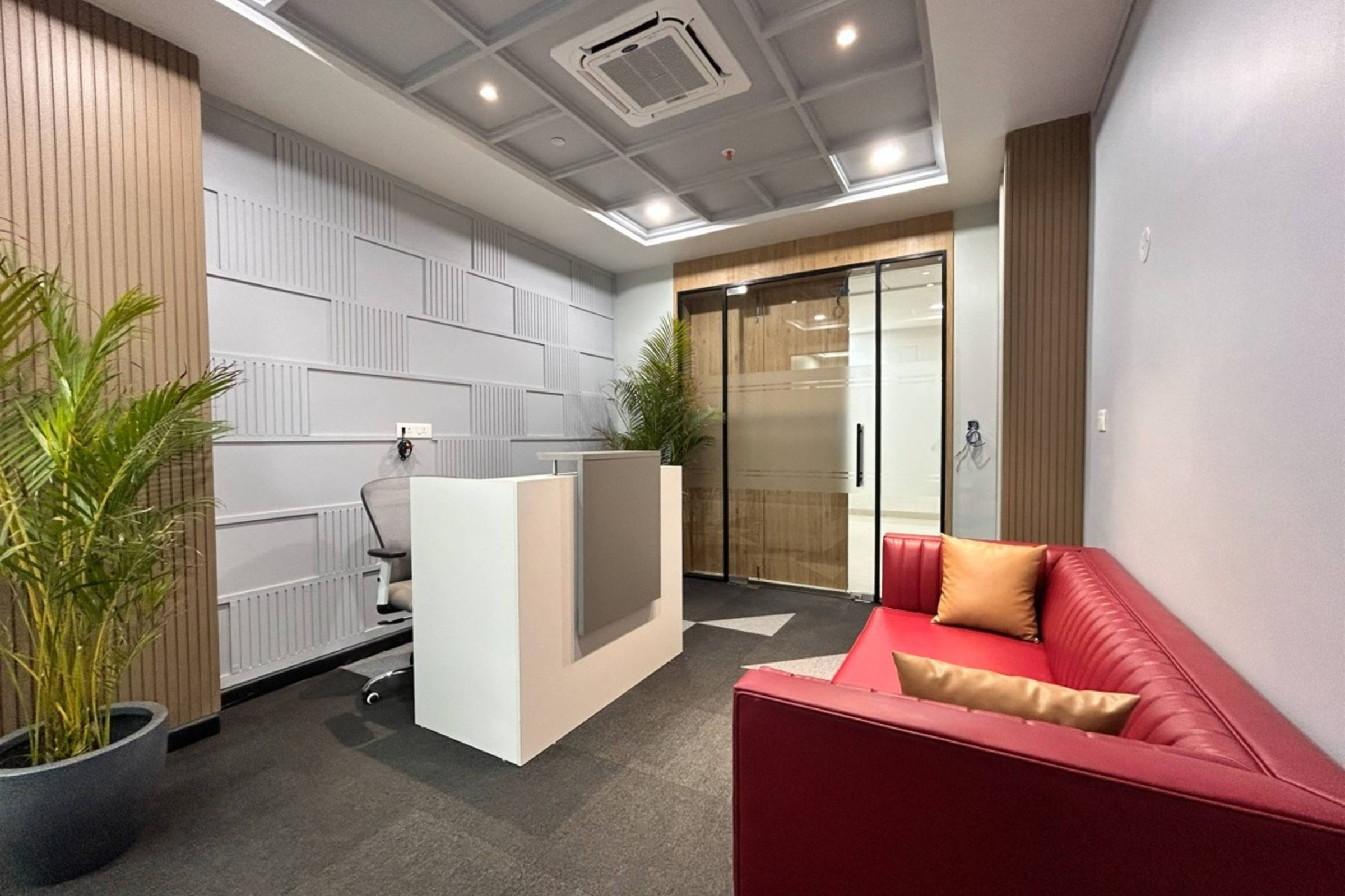 Modern office reception area with a white desk, gray office chair, glass door, potted plants, and a red sofa with gold cushions—all enhanced by fresh new construction painting.