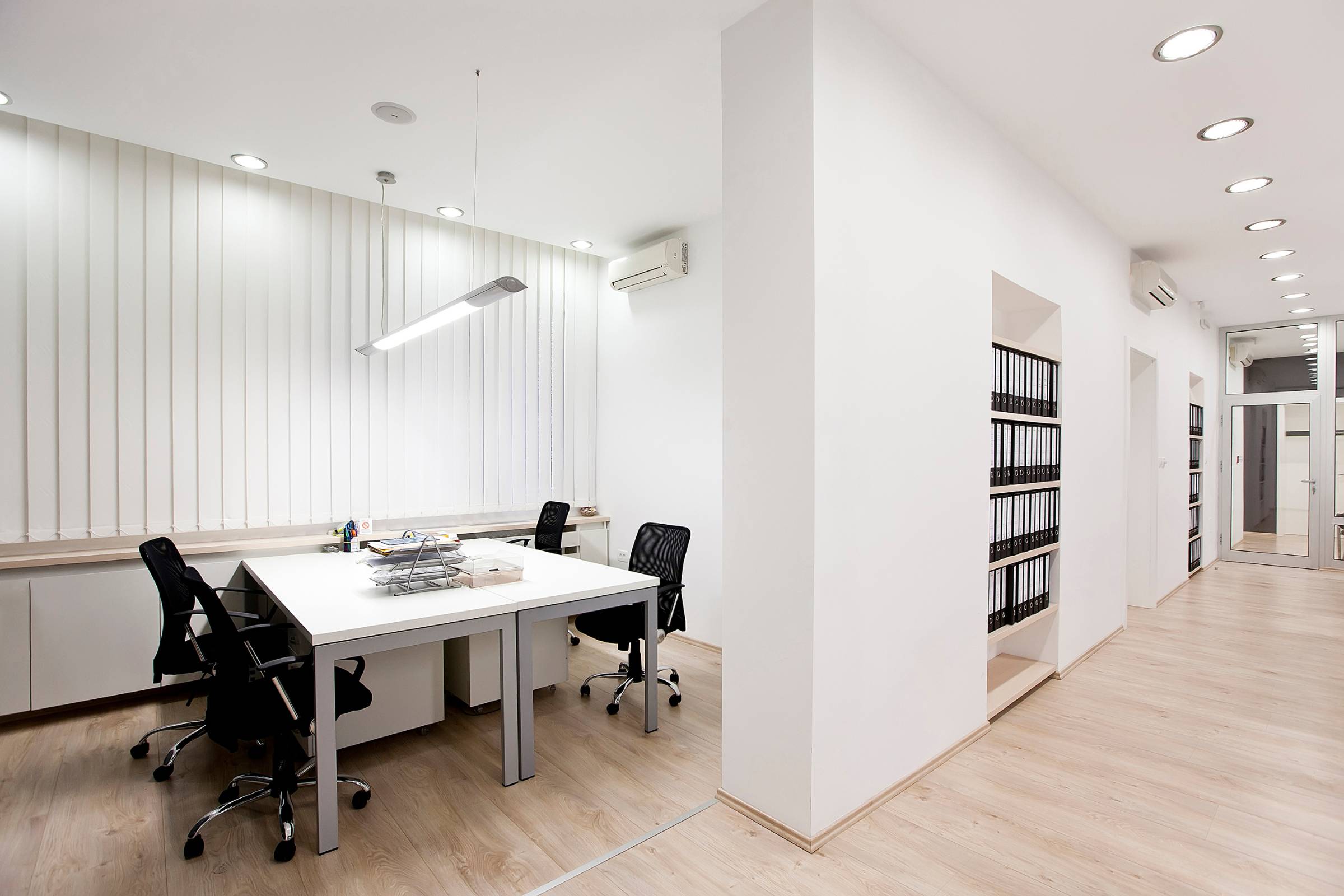 Modern office interior with white walls freshly completed by New Construction Painting, light wood flooring, a desk with four black chairs, and shelves filled with black binders. The space is well-lit with ceiling lights.