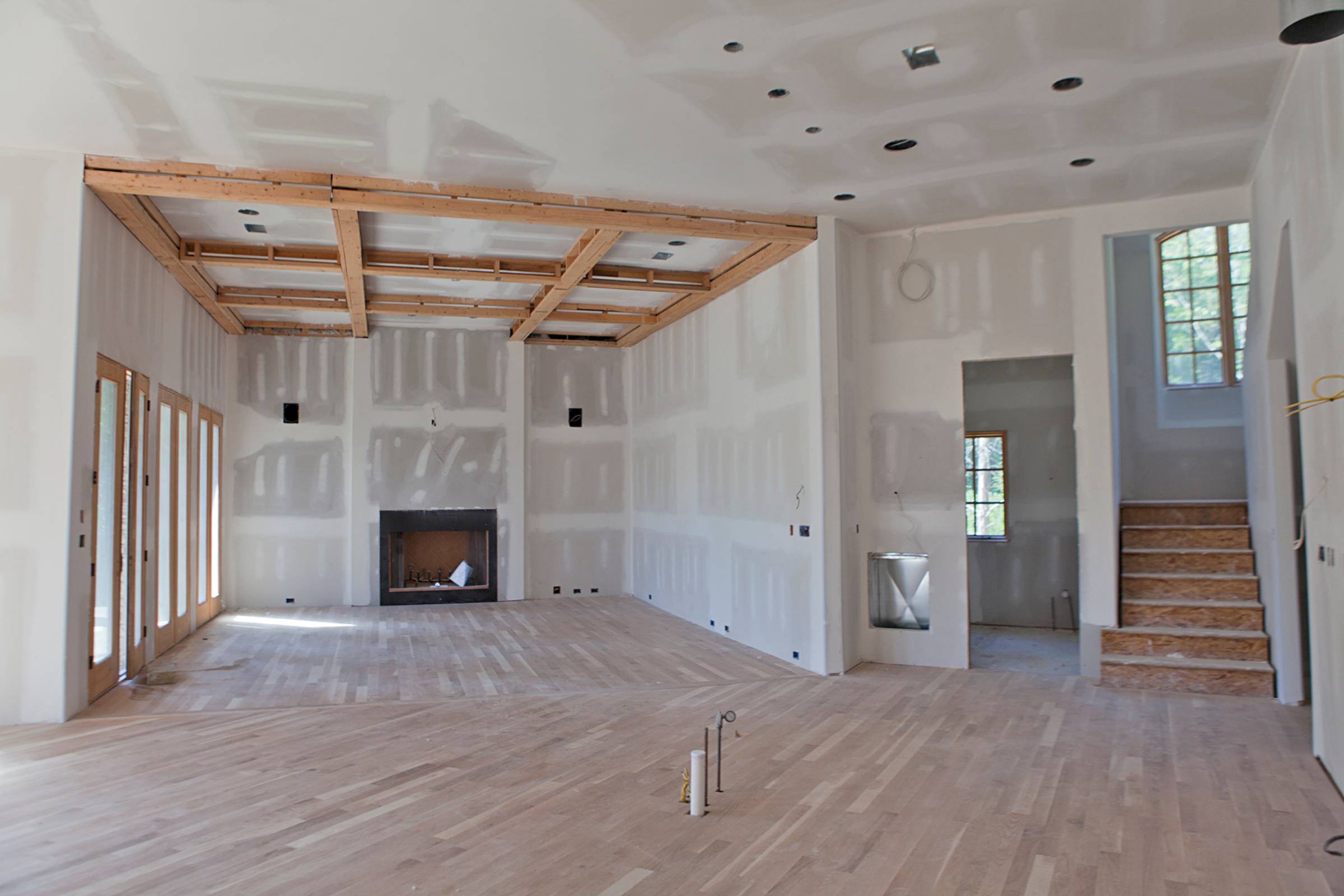 Unfinished living room with exposed drywall, wooden ceiling beams, fireplace, and hardwood floors; stairs and windows visible on the right side—ideal for New Construction Painting services.