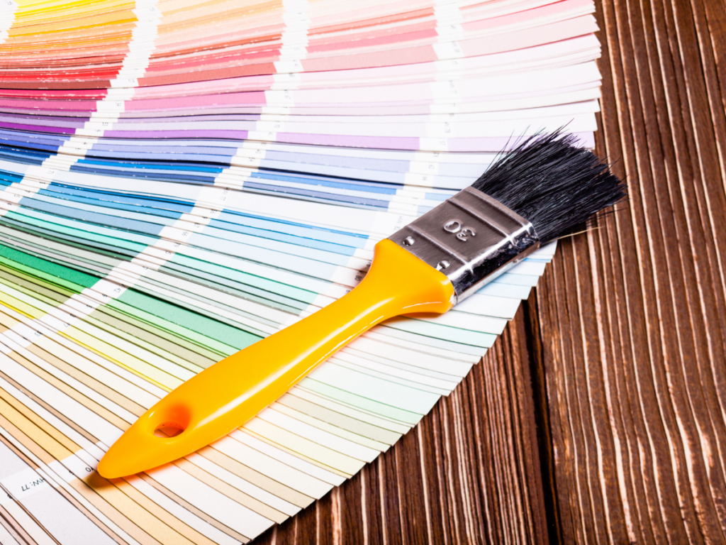 A paintbrush with an orange handle rests on a spread of paint color swatches arranged in a semicircle, ready to help you choose the perfect shade for your home on a wooden surface.