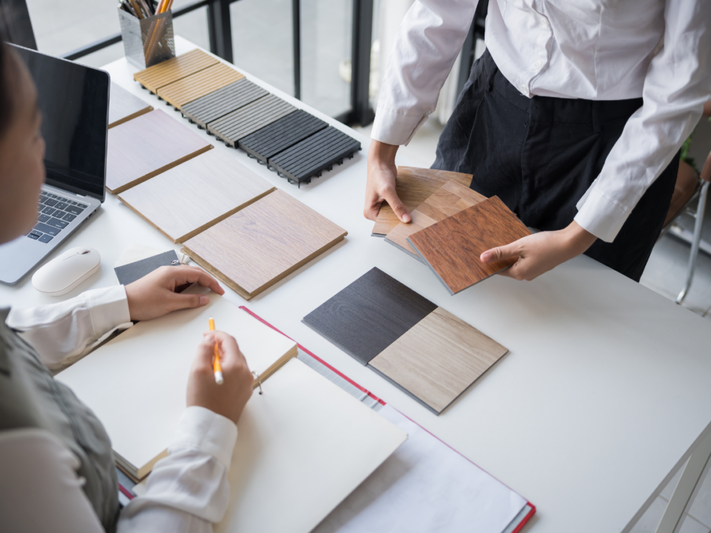 Two people review various wooden laminate samples on a desk with a laptop, notebook, and folders, likely discussing design or material selections as they choose finishes and paint colors for their home.