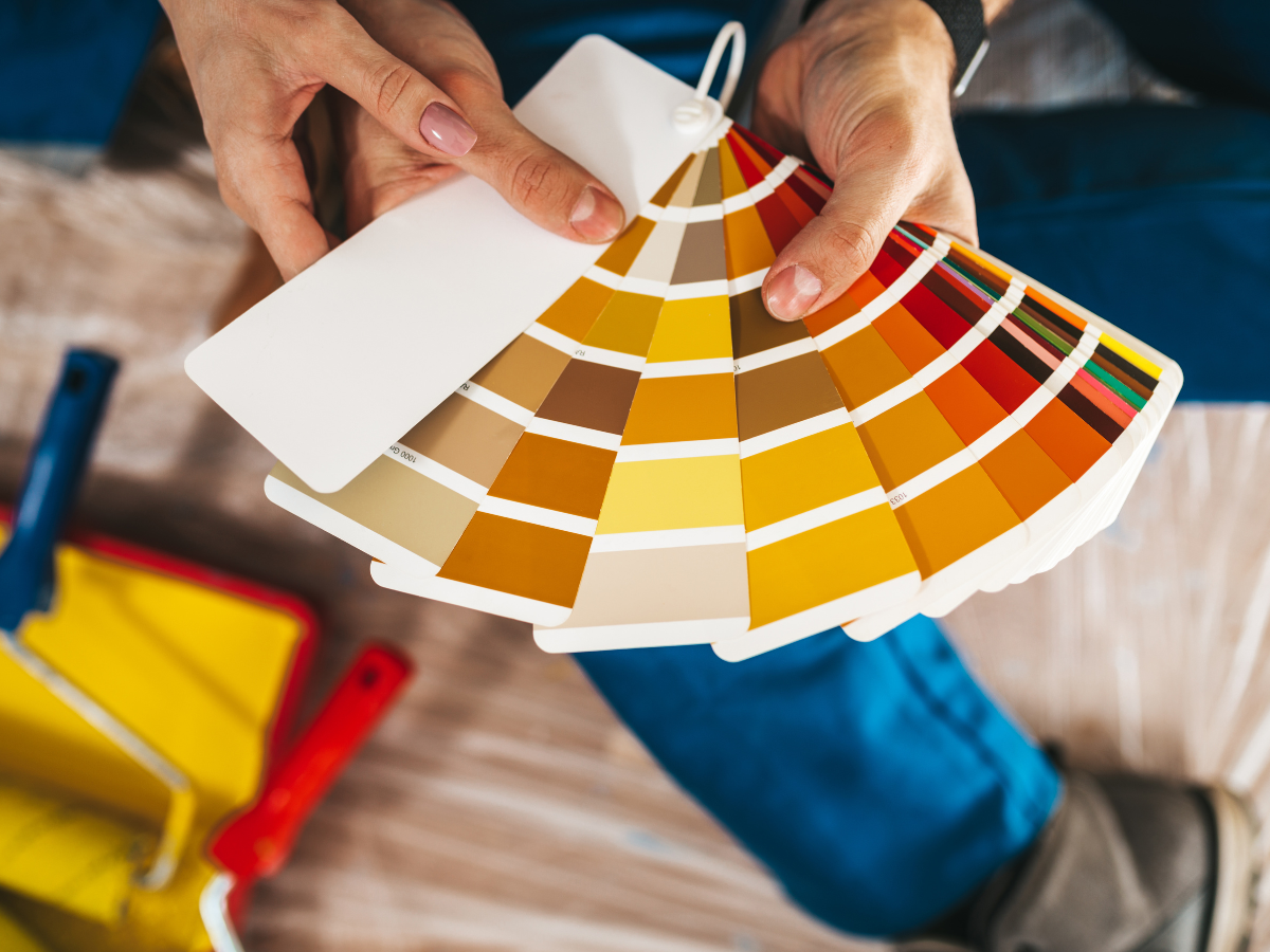 A person holds a color swatch book with shades of yellow, orange, and brown, demonstrating how to choose paint colors for your home, with paint rollers and trays in the background.