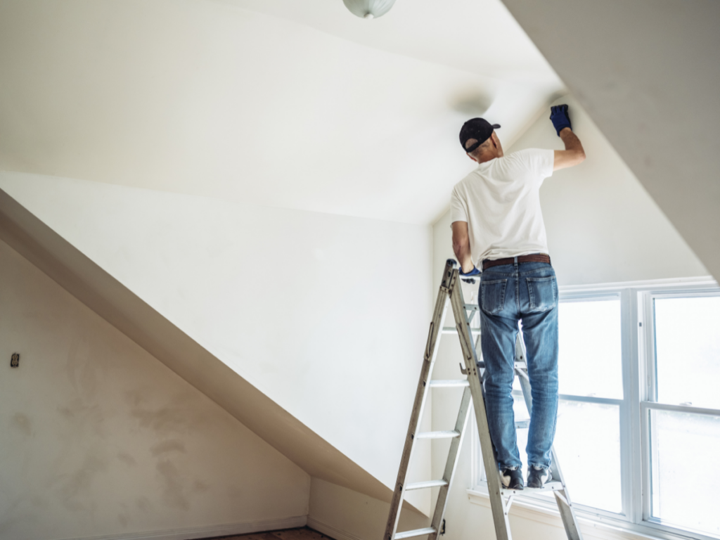 A painting contractor stands on a ladder, carefully painting the upper corner of a white wall in a room with slanted ceilings and a window in Marietta, GA.