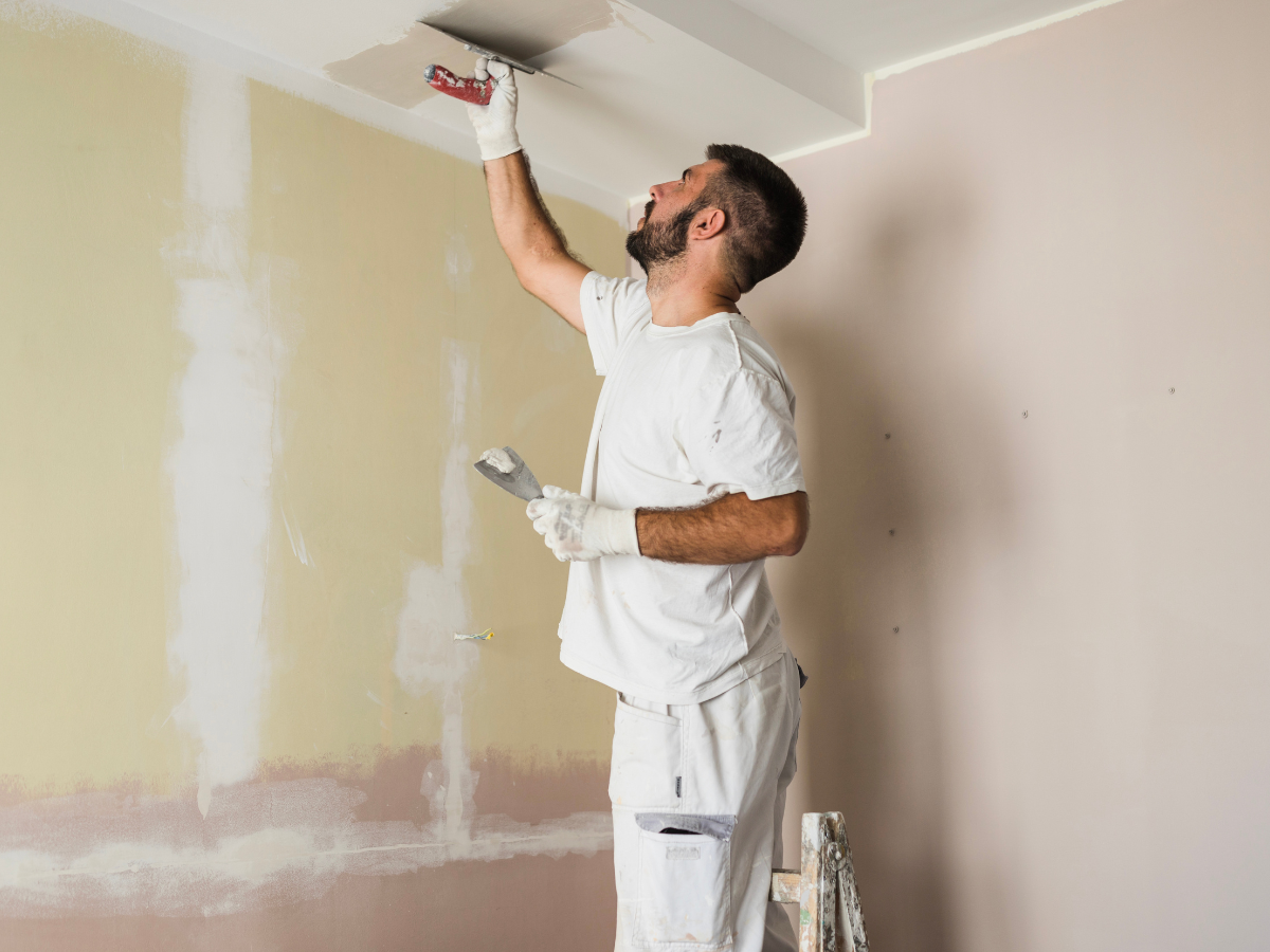 A painting contractor Marietta GA stands on a ladder, applying putty to the ceiling with a putty knife, preparing the wall for painting in a room under renovation.