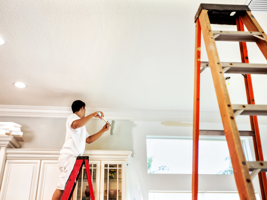 A person standing on a ladder paints the upper wall near the ceiling of a room, showcasing the careful work you can expect when you choose a painting contractor in Marietta GA; another ladder is visible in the foreground.