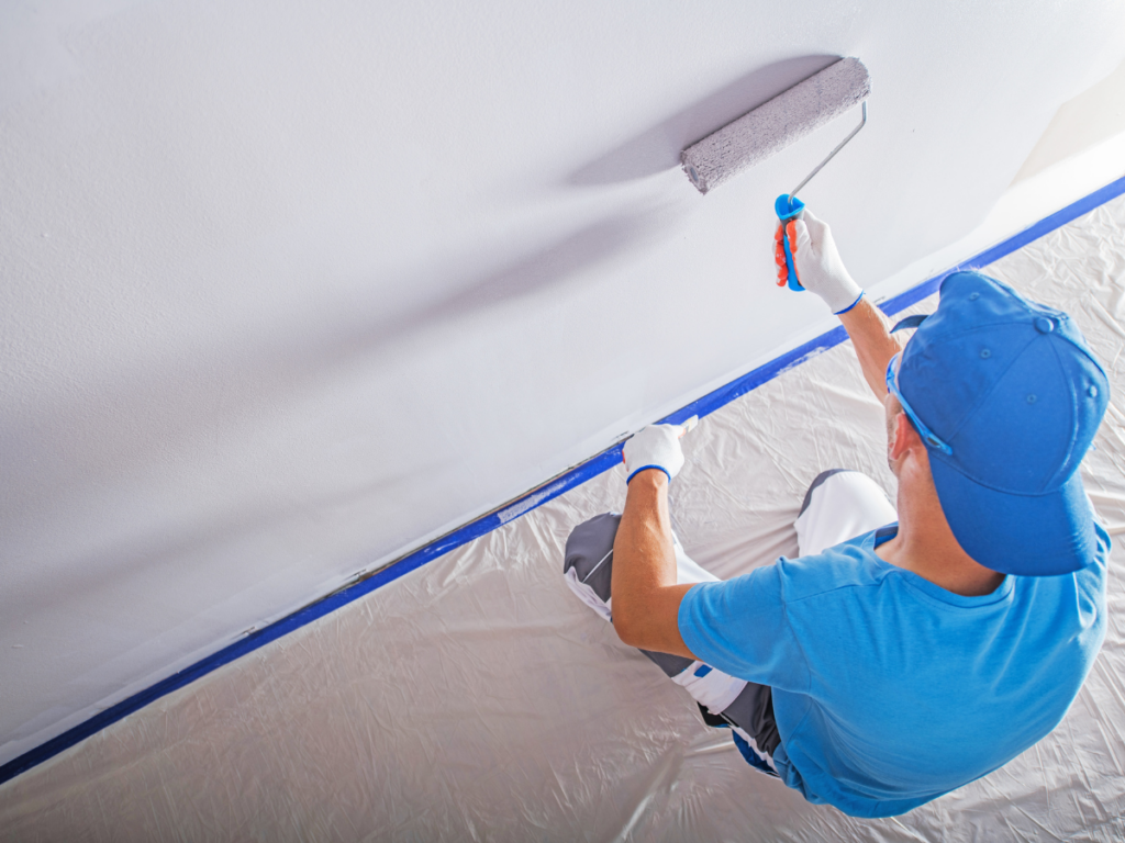 A painting contractor in Marietta GA, wearing a blue shirt and cap, uses a paint roller to paint a ceiling white while protective plastic covers the floor.