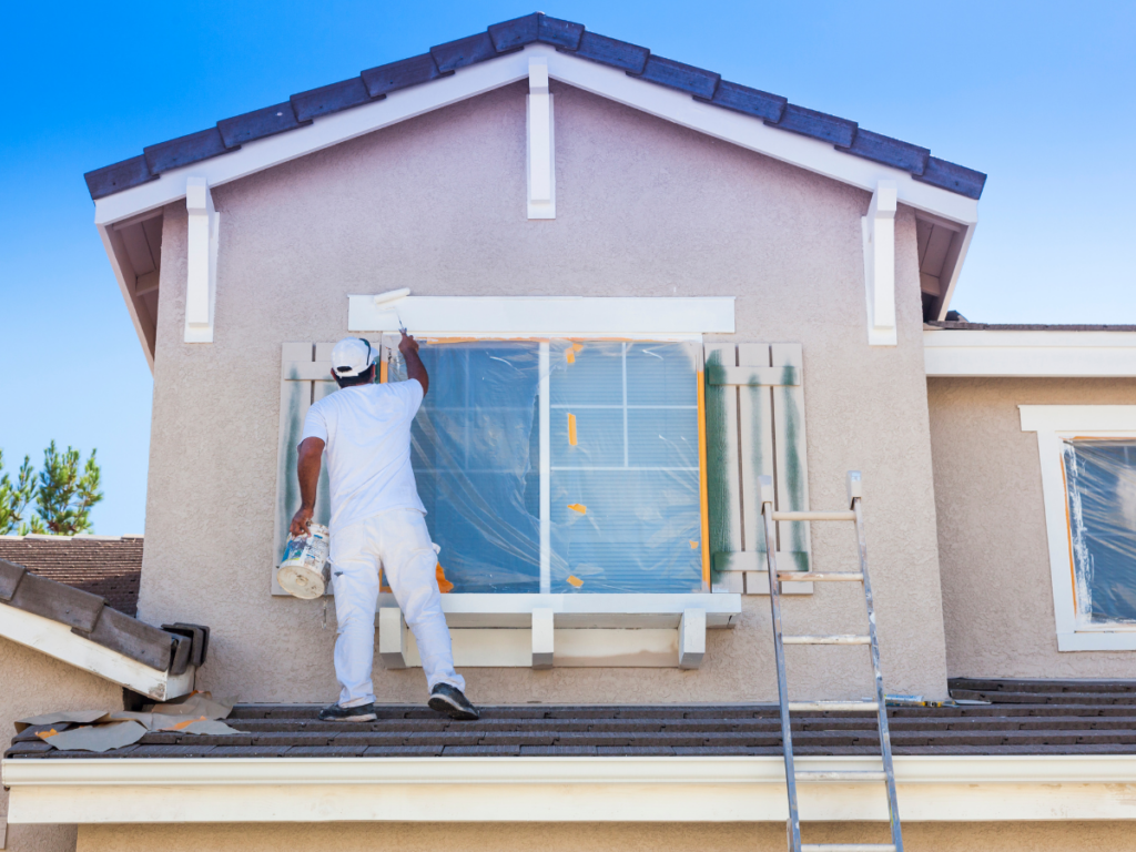 A person stands on a roof painting the exterior trim of a house window, protected by plastic sheeting—a perfect scene highlighting professional painting vs DIY painting. A ladder leans against the house for access.