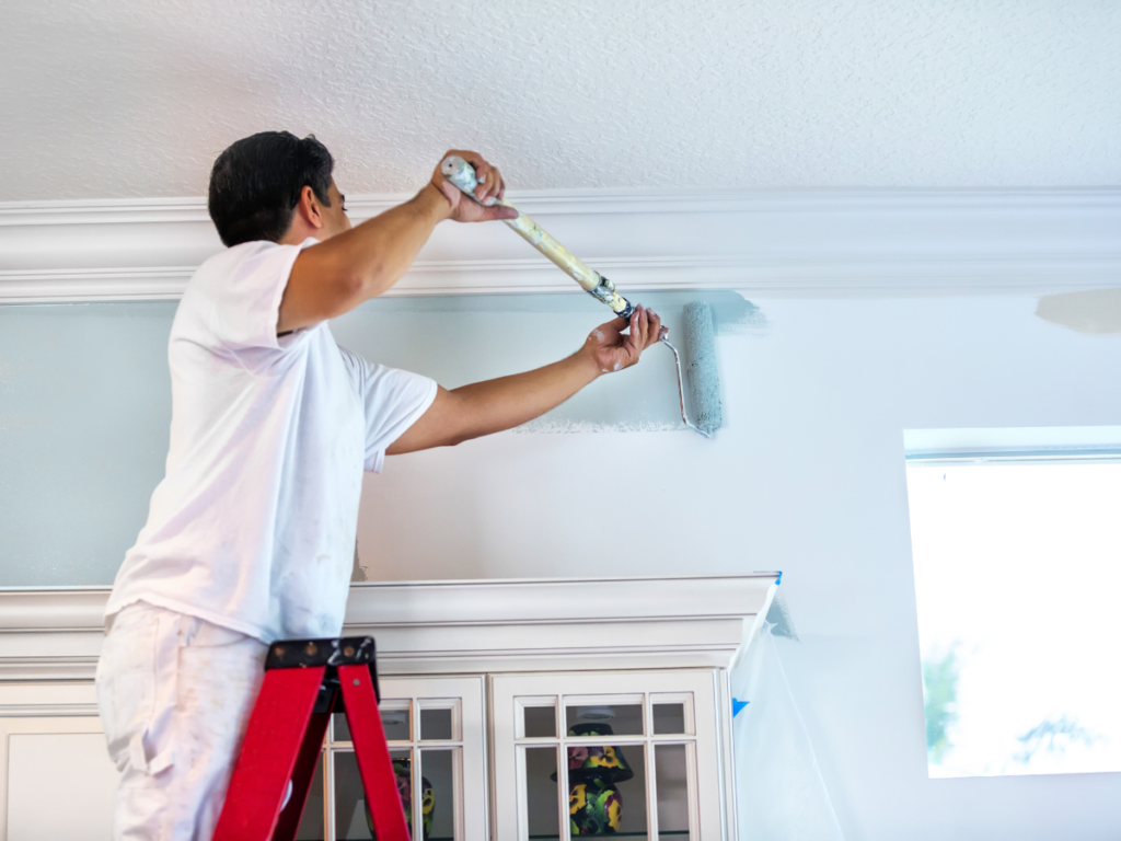 A person standing on a red ladder uses a paint roller to paint a light blue color on a wall near the ceiling above a white cabinet, showcasing the difference between professional painting vs DIY painting techniques.
