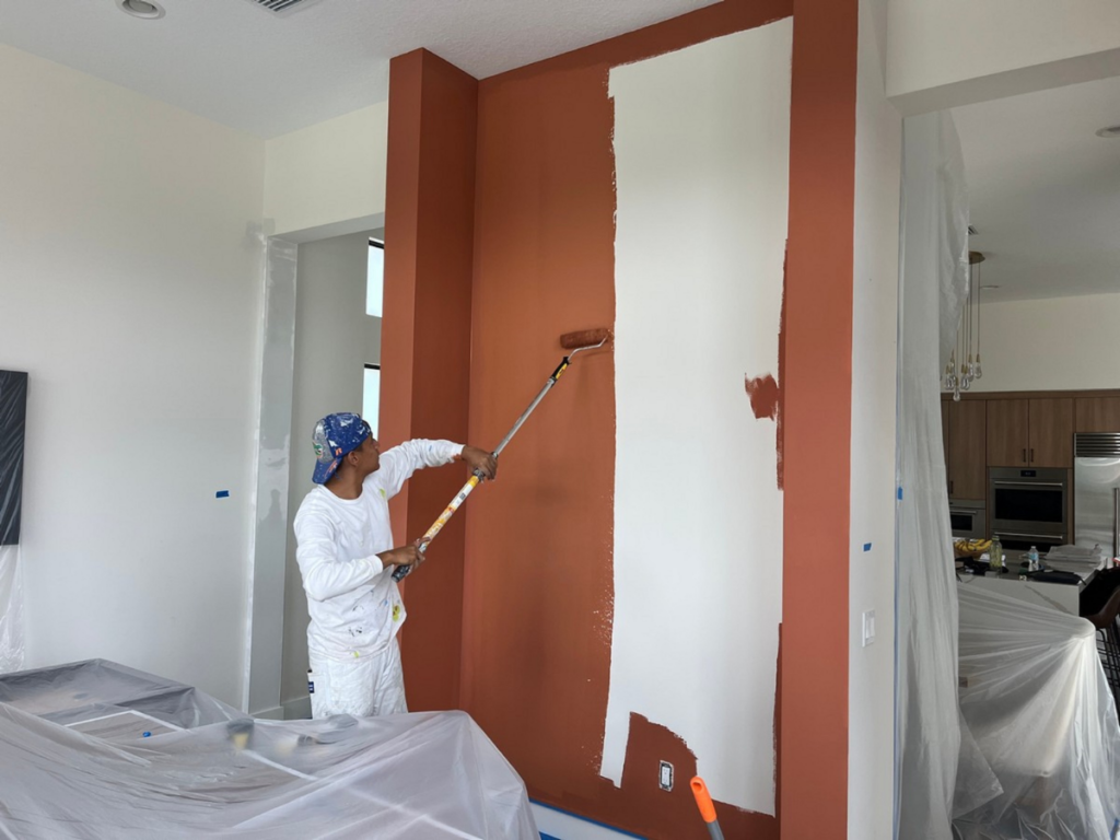 A person in white work clothes demonstrates professional painting vs DIY painting by expertly using a roller to paint a brown wall white, with all furniture and fixtures carefully covered for protection.
