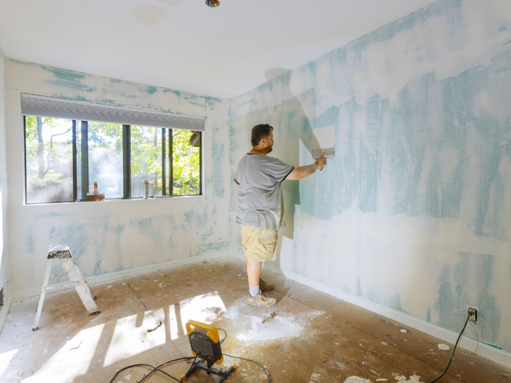 A person uses a roller to paint the wall of a partially renovated room with unfinished floors and patchy blue and white walls, illustrating the difference between professional painting vs DIY painting.