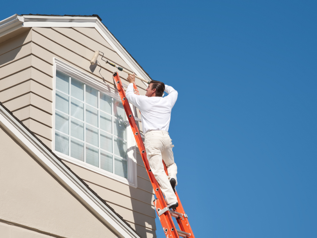 A person on an orange ladder paints the exterior wall of a house with a roller brush near a second-story window under a clear blue sky, highlighting the difference between professional painting vs DIY painting.