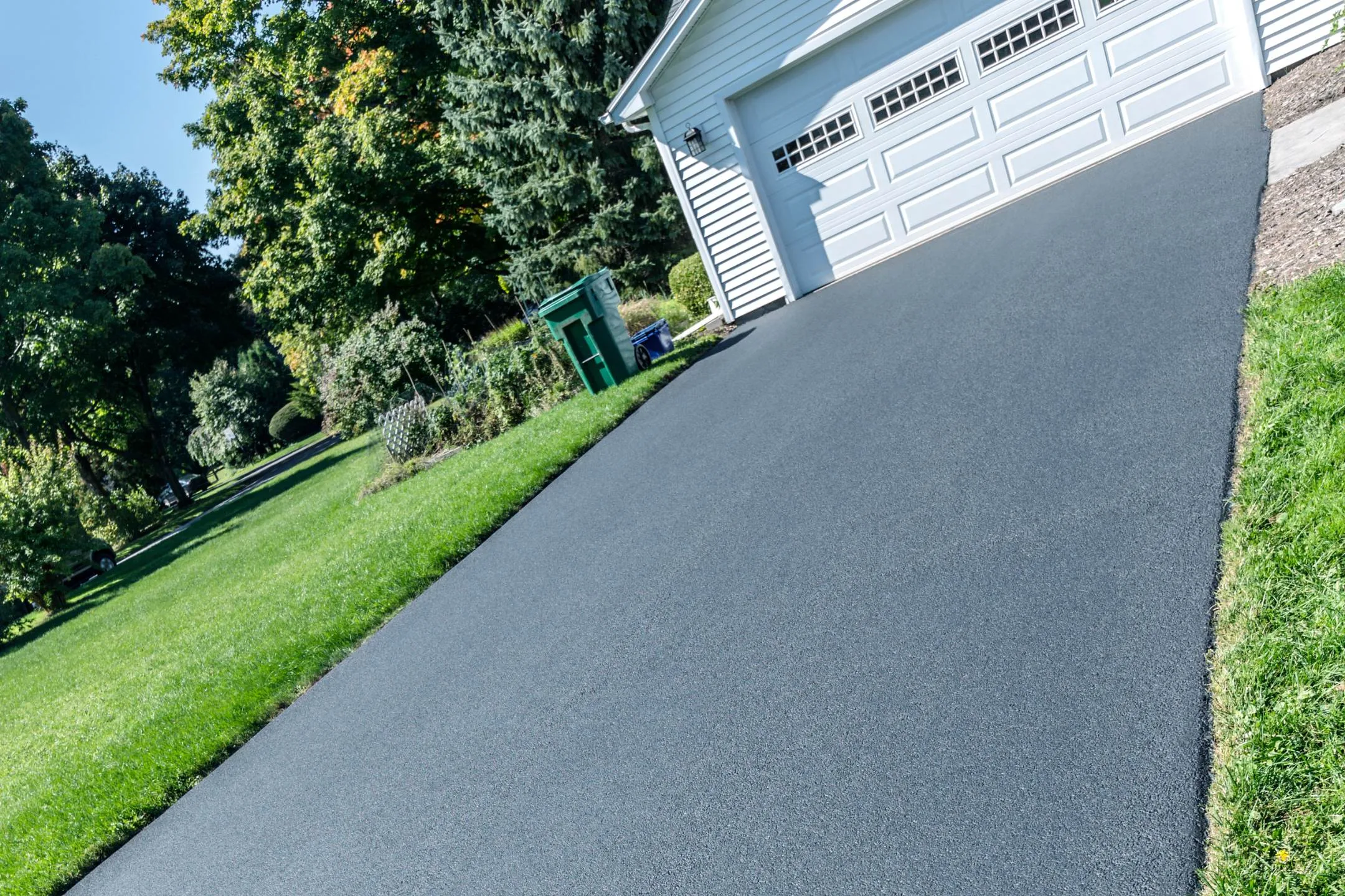 Freshly paved asphalt driveway leading to a white garage with three doors, enhanced with residential epoxy coatings, surrounded by green grass and trees on a sunny day.