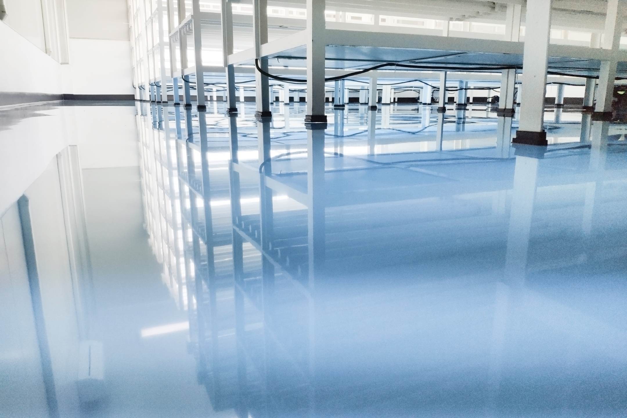 A low-angle view of a clean, shiny floor finished with Residential Epoxy Coatings reflects white metal shelving units and bright windows in an industrial or laboratory setting.