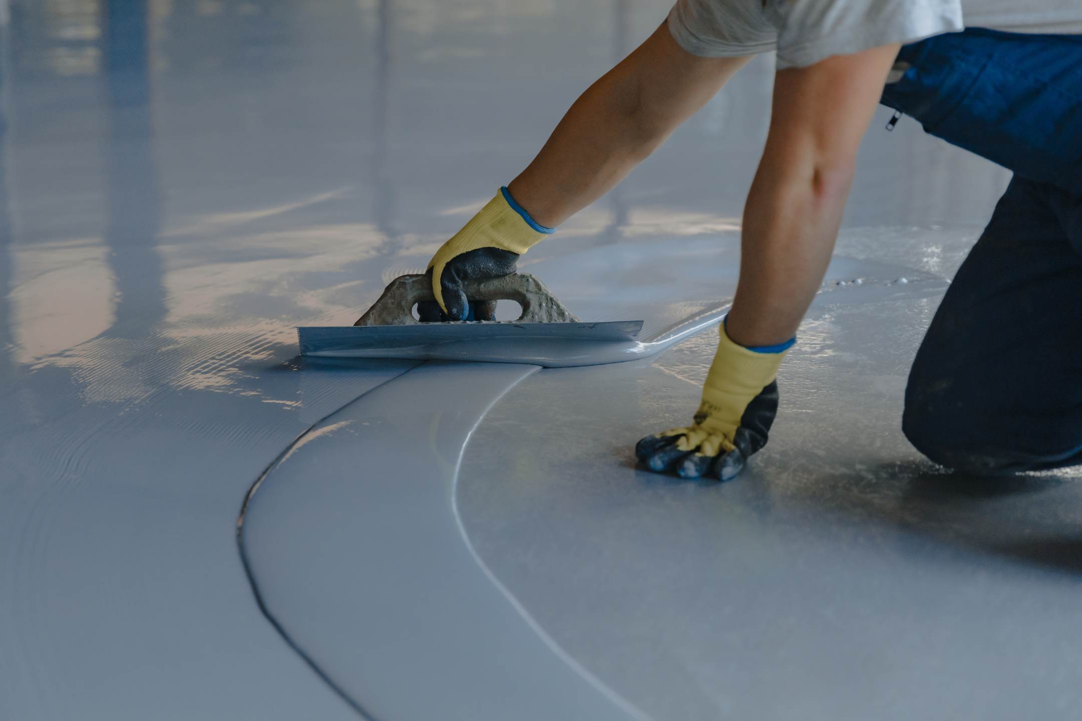 A person wearing gloves uses a trowel to spread a smooth layer of gray Residential Epoxy Coatings on a floor.