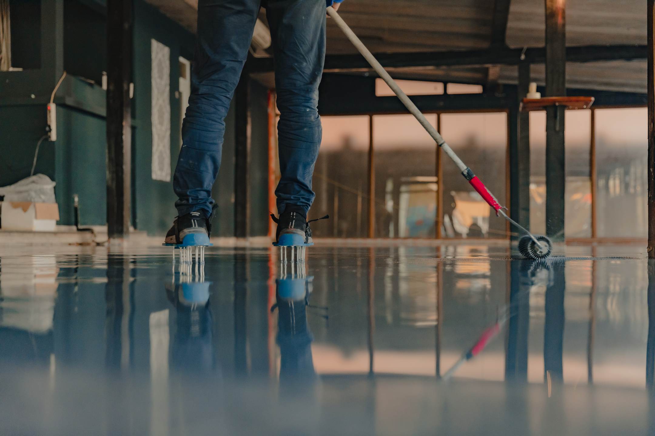 Wearing spike shoes, a person uses a roller tool to smooth Residential Epoxy Coatings on a reflective floor in an unfinished indoor space.