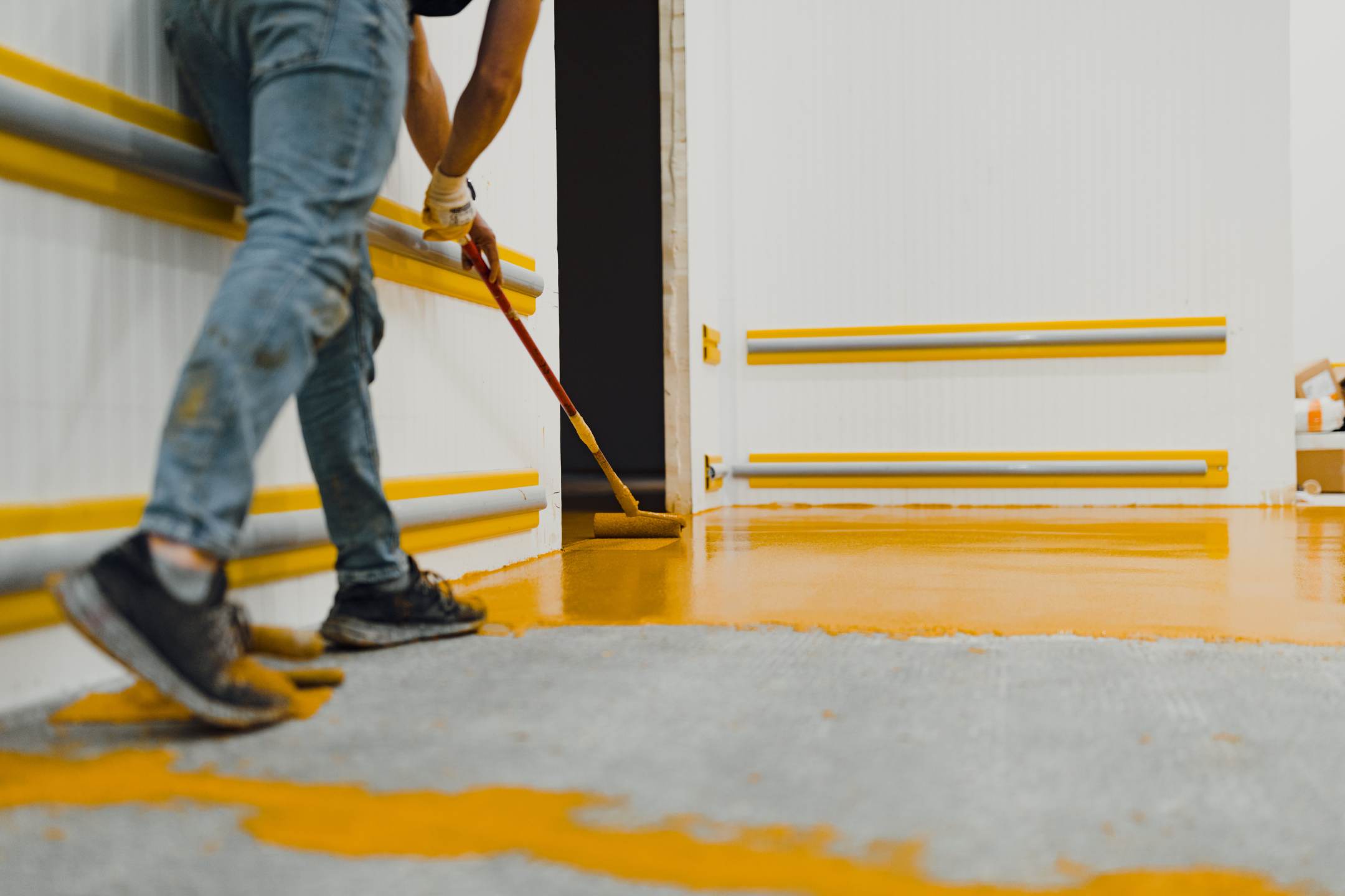 Person using a roller brush to apply yellow residential epoxy coatings to a floor in a room with white walls and yellow railings.