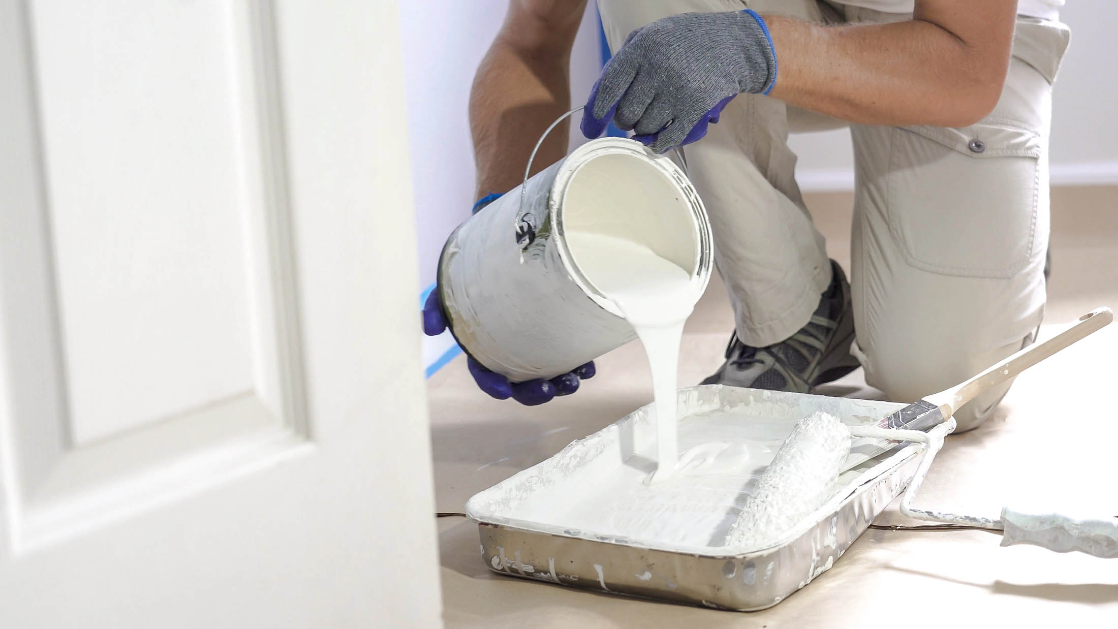 Person wearing gloves pours white paint from a can into a paint tray, preparing to use a roller for New Construction Painting indoors.