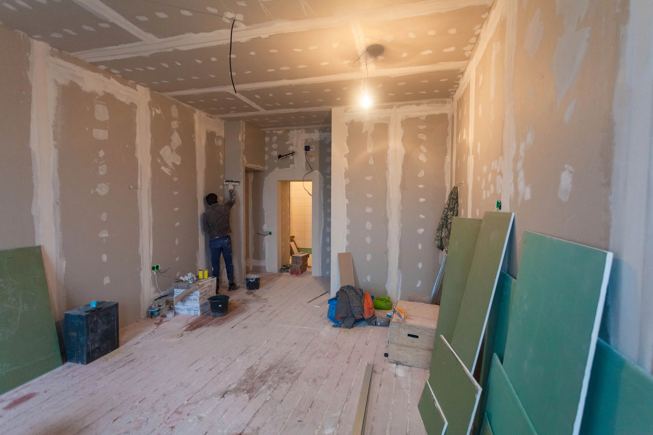 A person is working on renovating a room with unfinished drywall and exposed wiring, preparing the space for New Construction Painting services amid scattered construction materials.