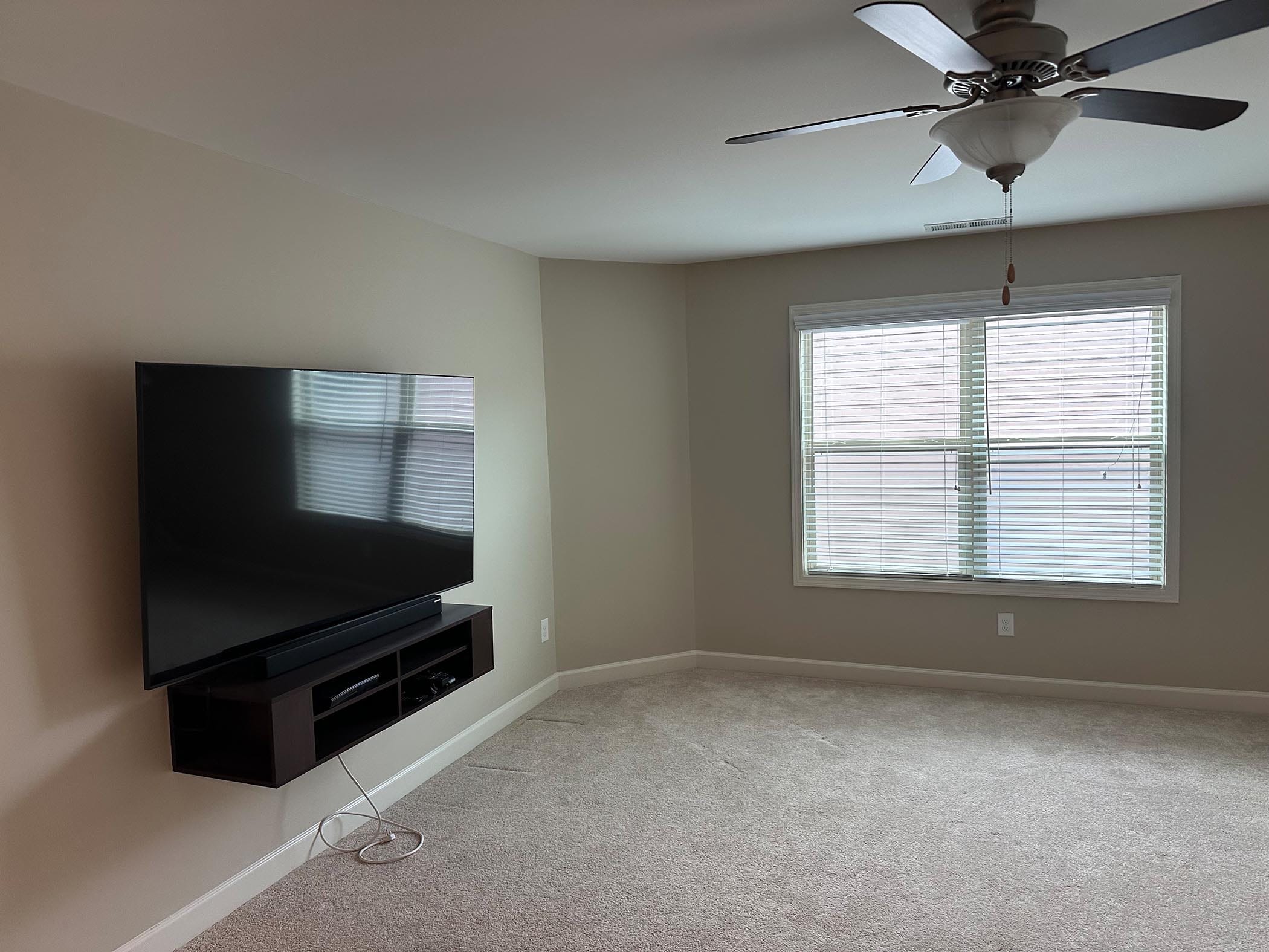 A carpeted living room with a wall-mounted TV, a small media shelf, a window with blinds, and a ceiling fan with light.