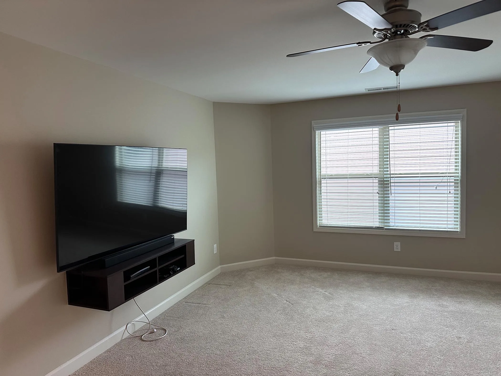 A carpeted living room with a wall-mounted TV, a small media shelf, a window with blinds, and a ceiling fan with light.