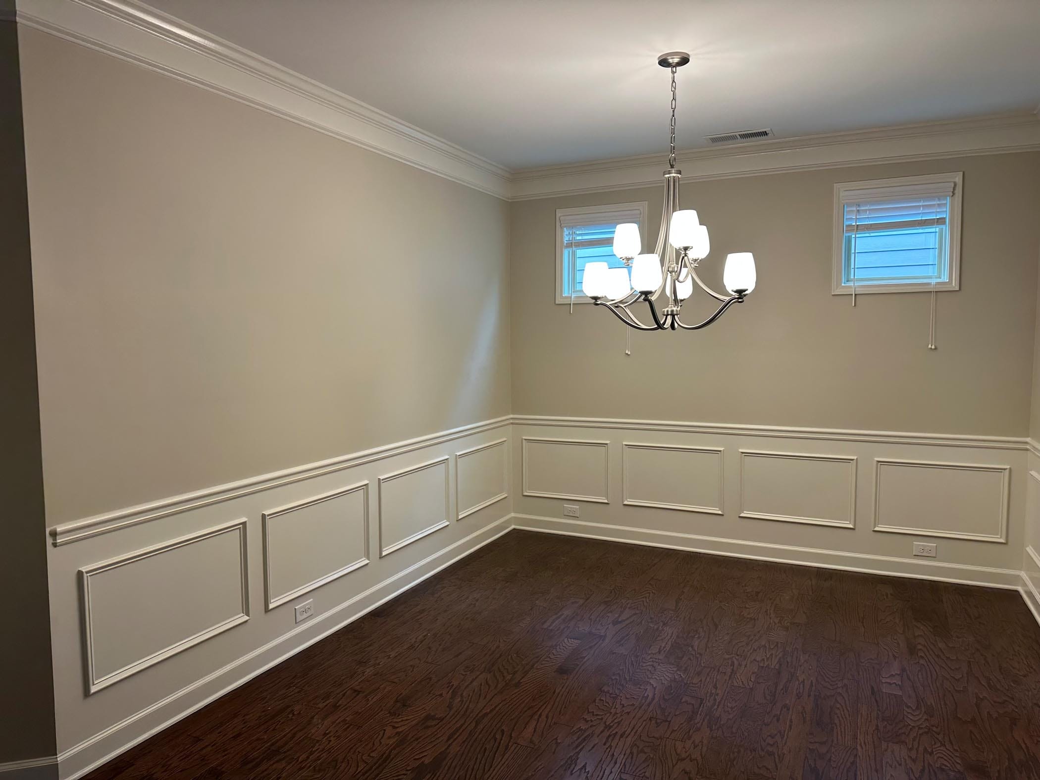 Empty dining room with beige walls, dark wood floor, chandelier light fixture, two small windows, and decorative wall trim.