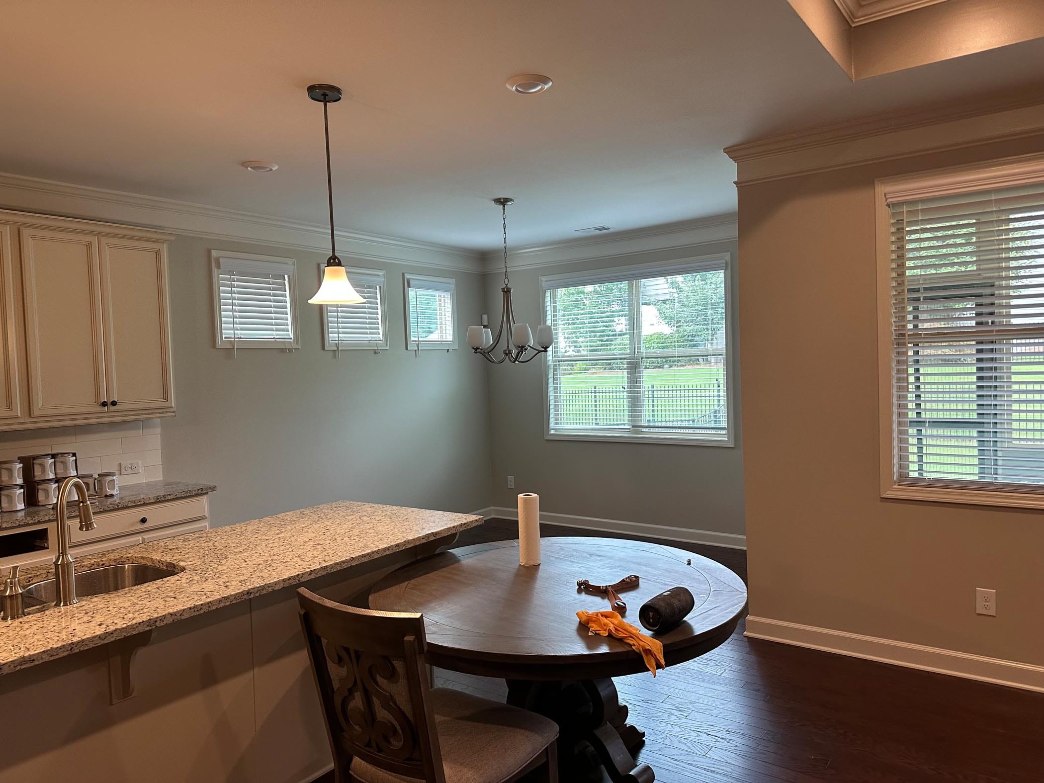 A kitchen and dining area with granite counters, a round table, chairs, pendant and chandelier lighting, and windows overlooking a grassy yard. A paper towel roll and cleaning items are on the table.