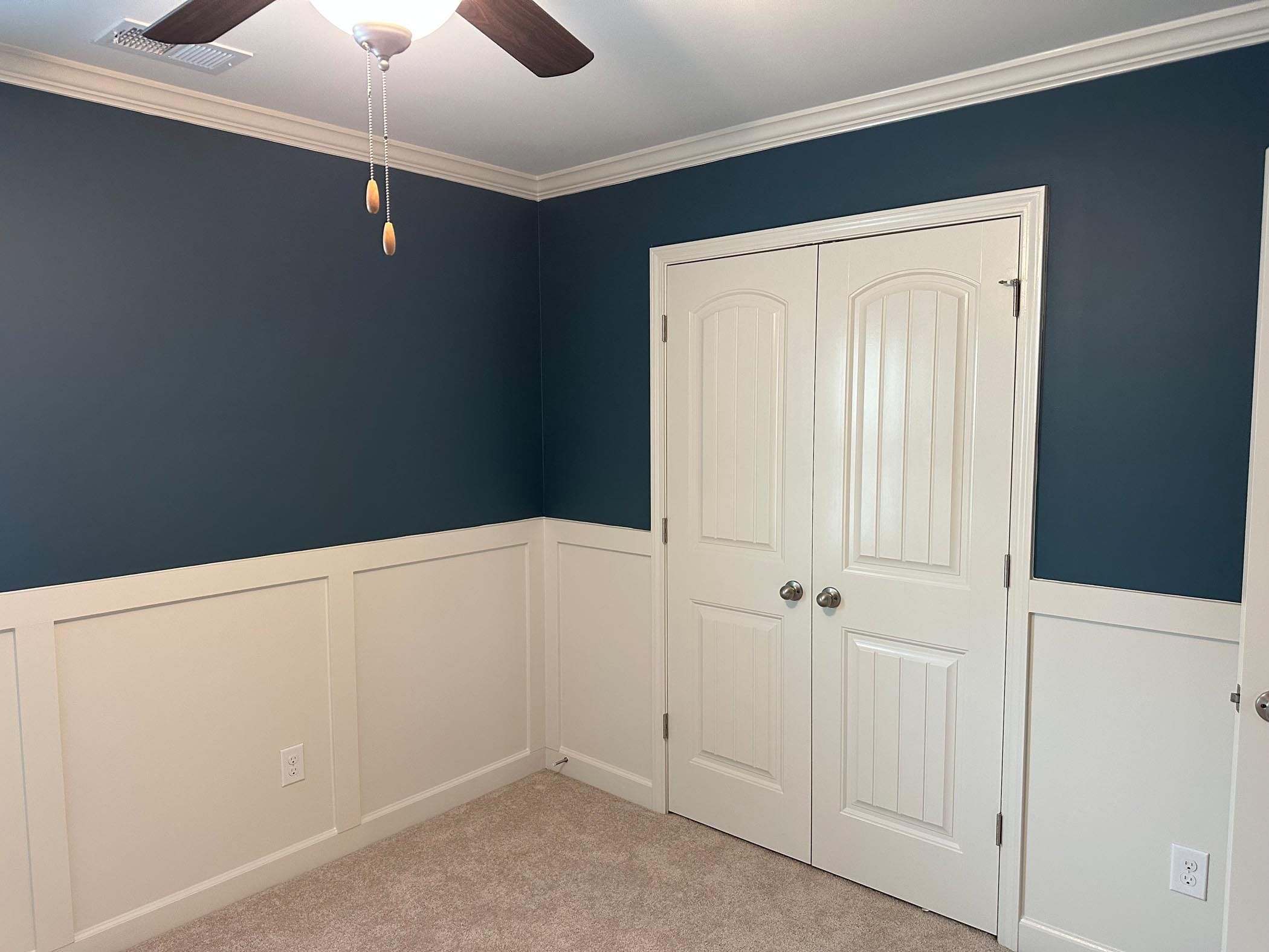 Room with blue upper walls, white wainscoting, beige carpet, a ceiling fan, and double white doors with silver knobs. A light switch and outlet are visible on the walls.