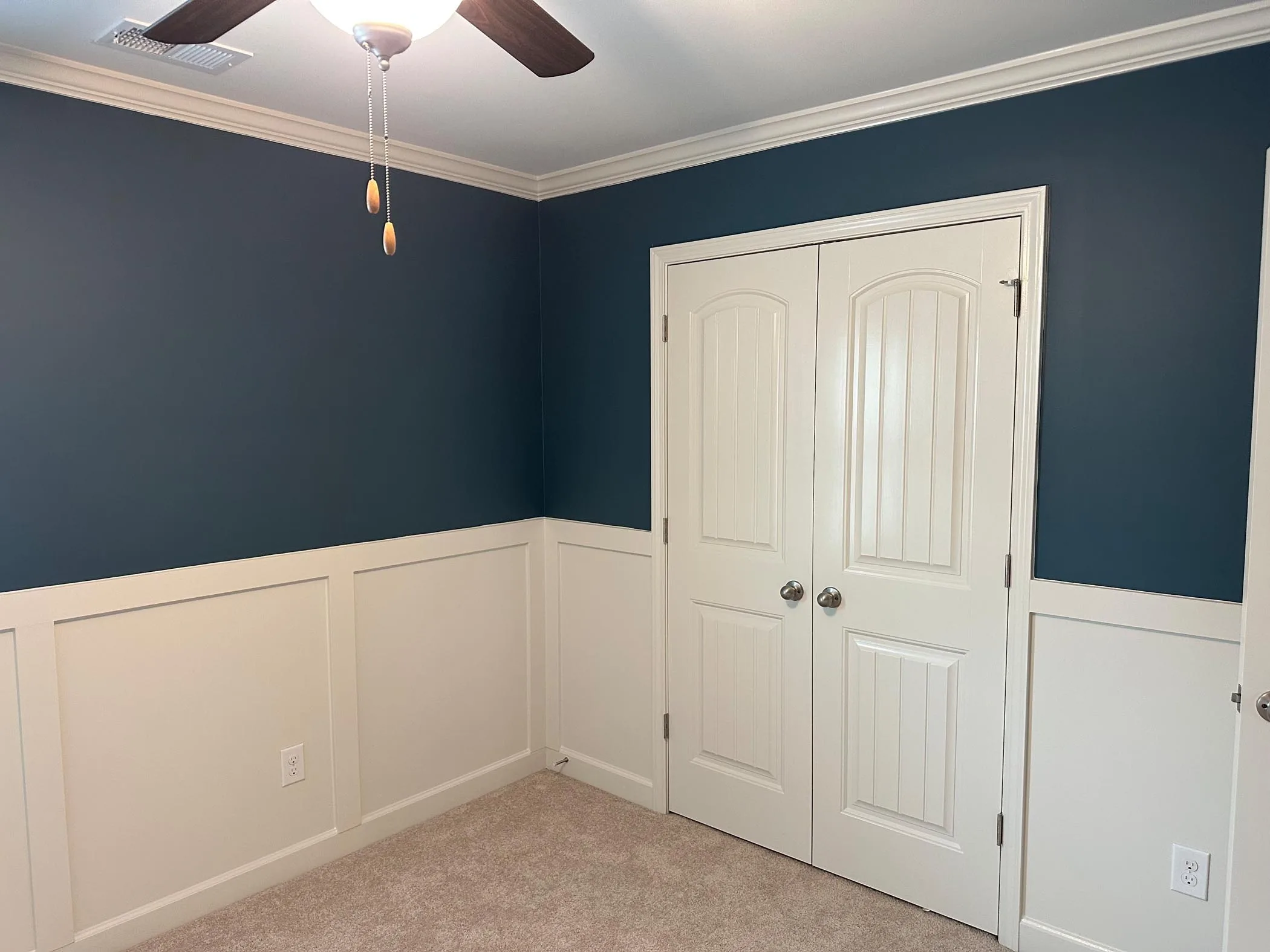 Room with blue upper walls, white wainscoting, beige carpet, a ceiling fan, and double white doors with silver knobs. A light switch and outlet are visible on the walls.