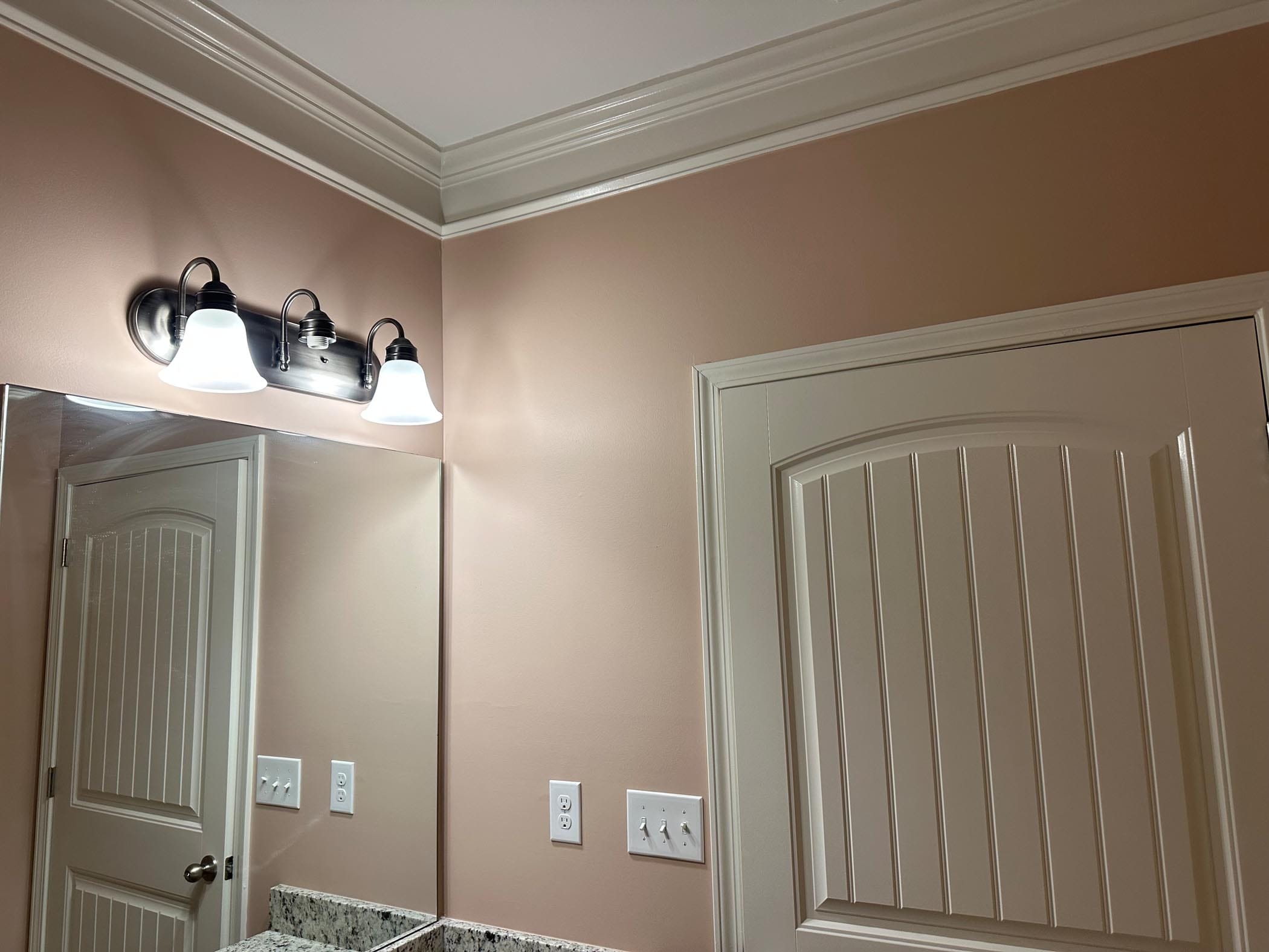 A bathroom corner with beige walls, a mirror, two light fixtures above it, and closed paneled doors. Several light switches and outlets are visible.