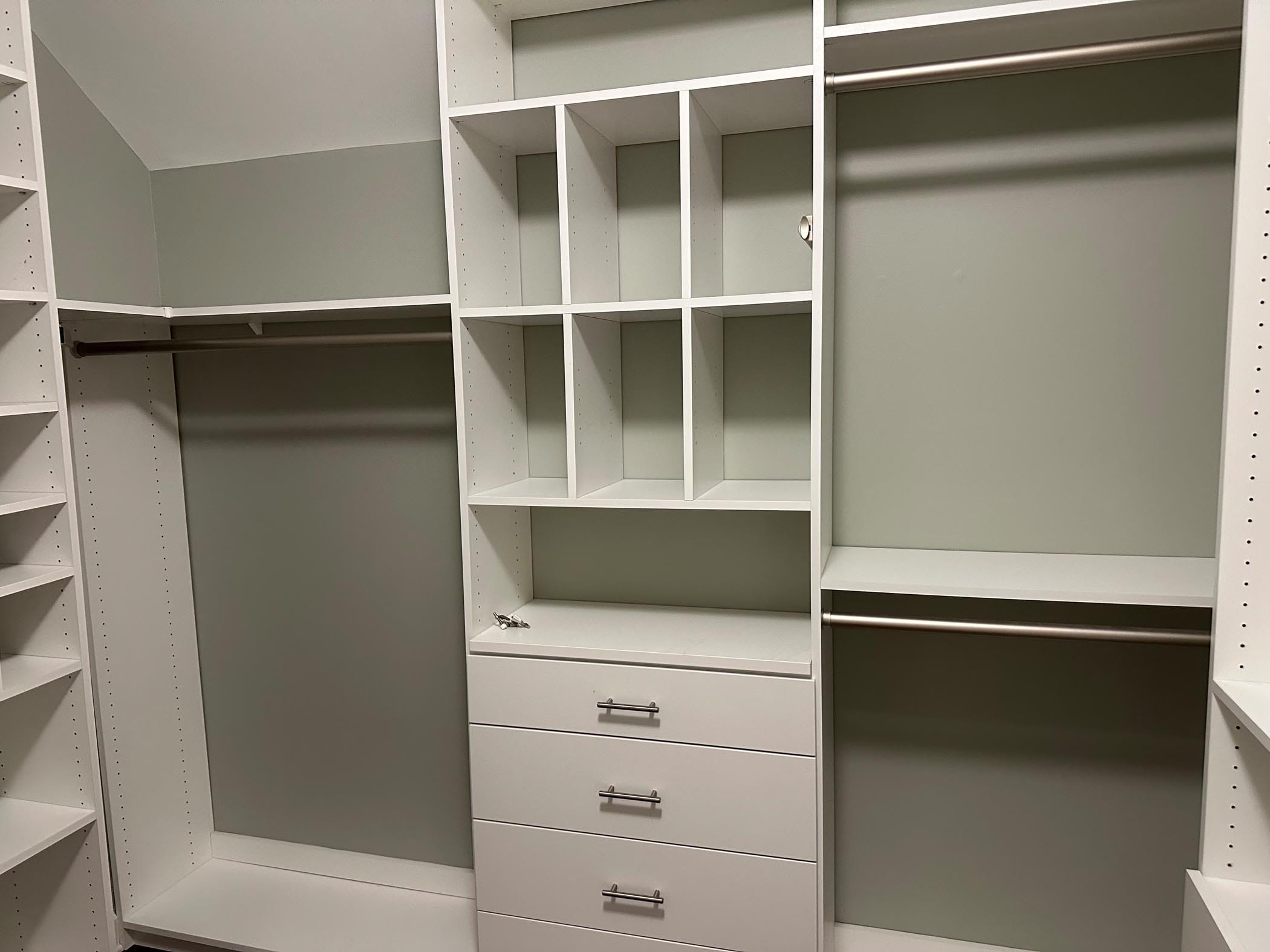White built-in closet organizer with open shelves, three drawers, and multiple hanging rods, installed against green walls in an empty walk-in closet.