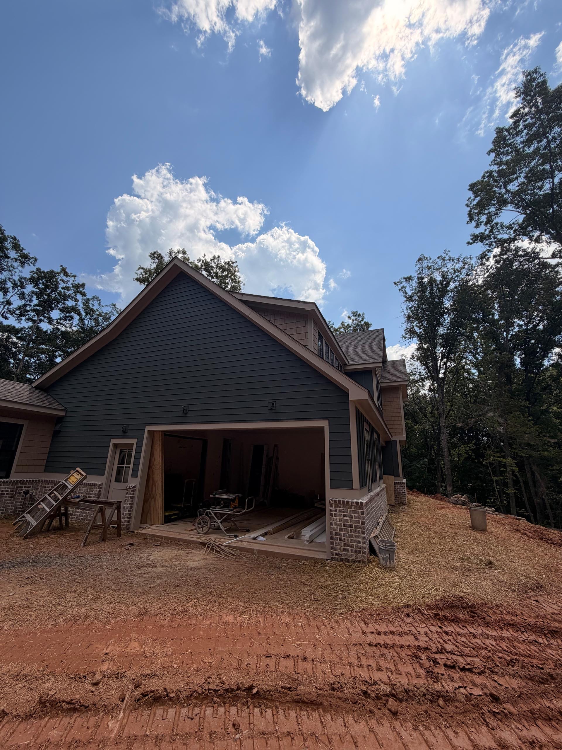A partially constructed house with an open garage, building materials, and tools scattered outside on a dirt lot under a partly cloudy sky.