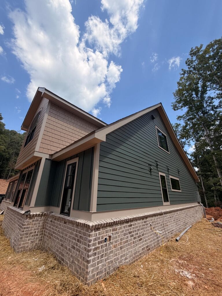 A newly constructed house with green siding, beige trim, and brick foundation under a partly cloudy sky.